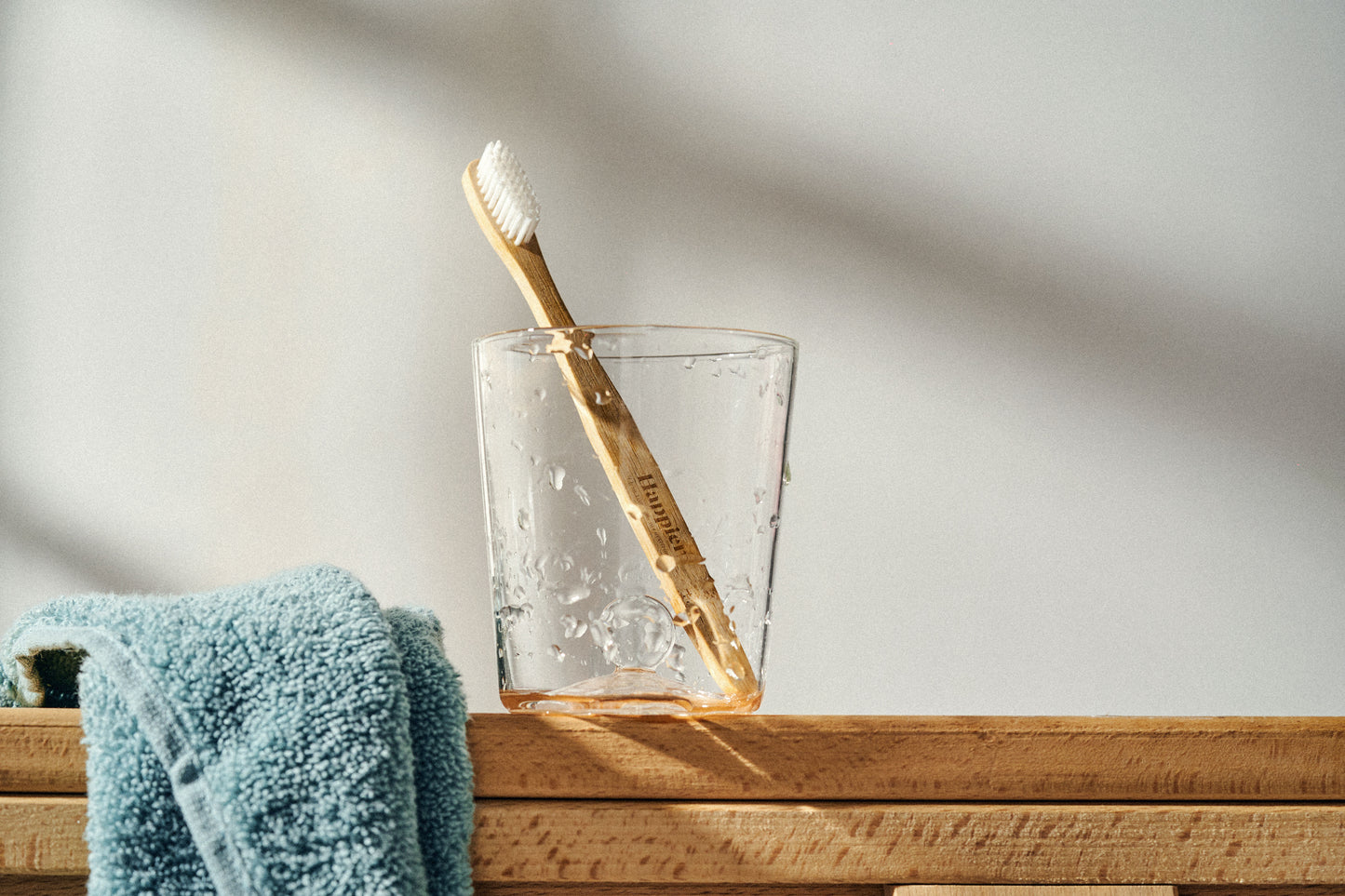 bamboo toothbrush in a clear glass tumbler sitting on a wooden surface with a turquoise fluffy towel next to it 