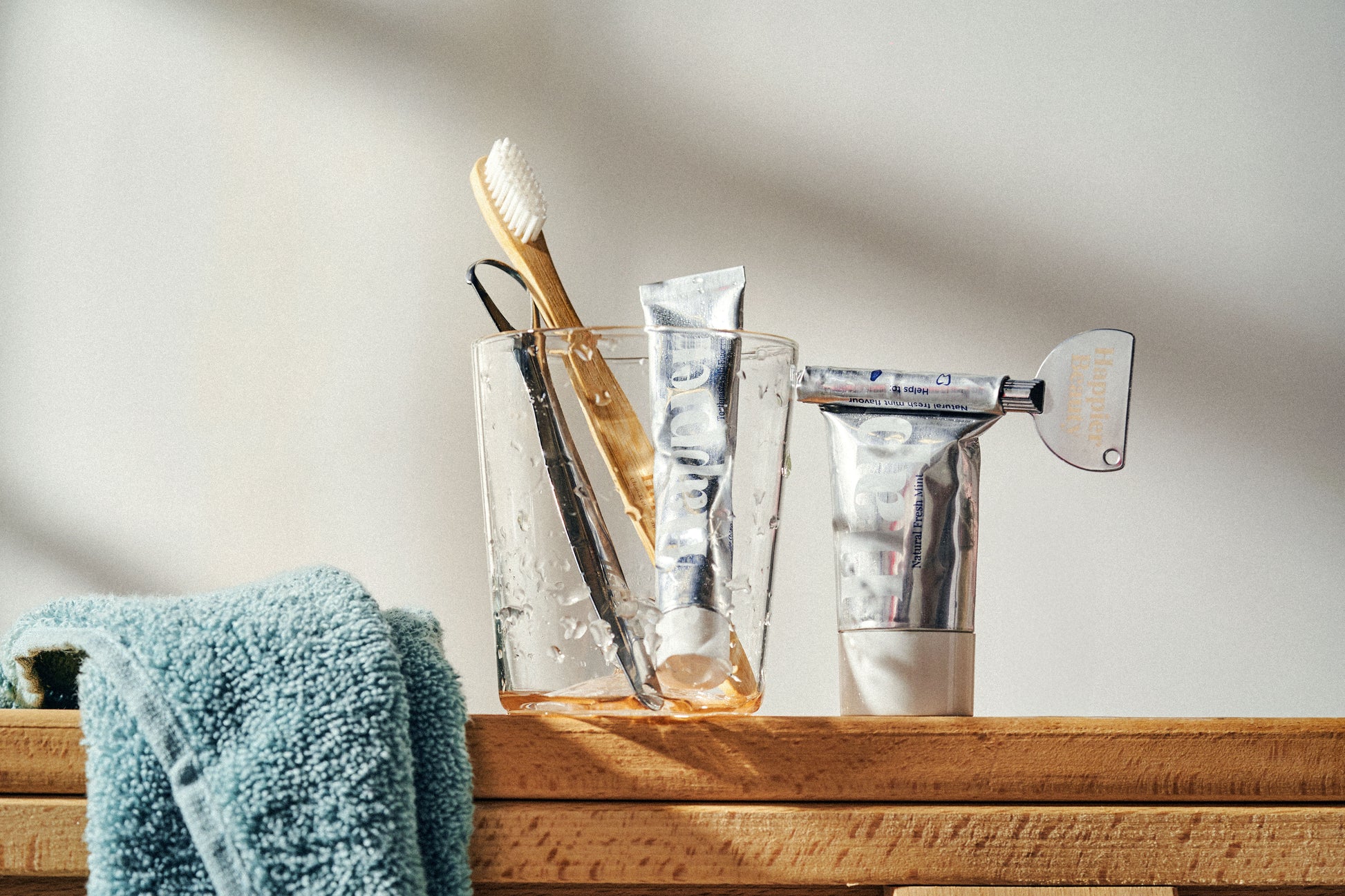 bamboo toothbrush, travel and full sized toothpaste in an aluminium tube and a stainless steel tongue scraper from happier beauty sitting in a clear glass on a wooden counter with a turquoise/dark blue fluffy towel draped over the wood