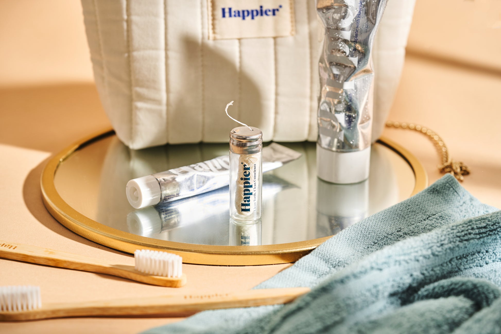 natural dental products from happier beauty on a peach background and sitting on top of a round mirror with gold edging and a turquoise fluffy towel. You can see the washbag, aluminium tubes, dental floss and bamboo toothbrushes