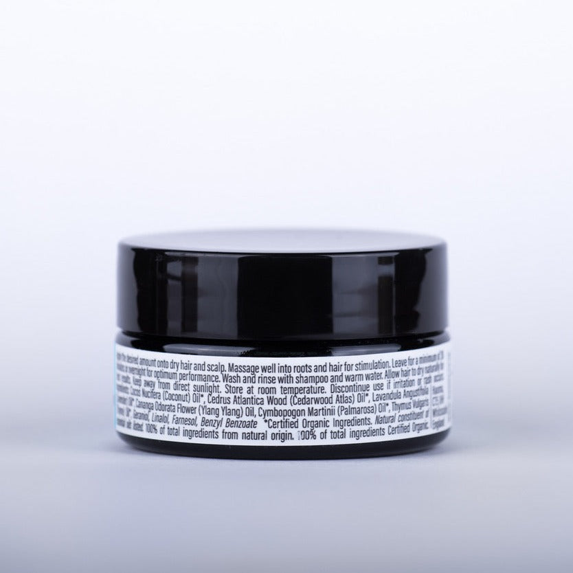 Photo of Haoma hair revitaliser form the back. You can see a black glass jar with black plastic lid which are equally sized. the glass is labelled with a white label which gives you directions for use and the full list of ingredients (INCI). 