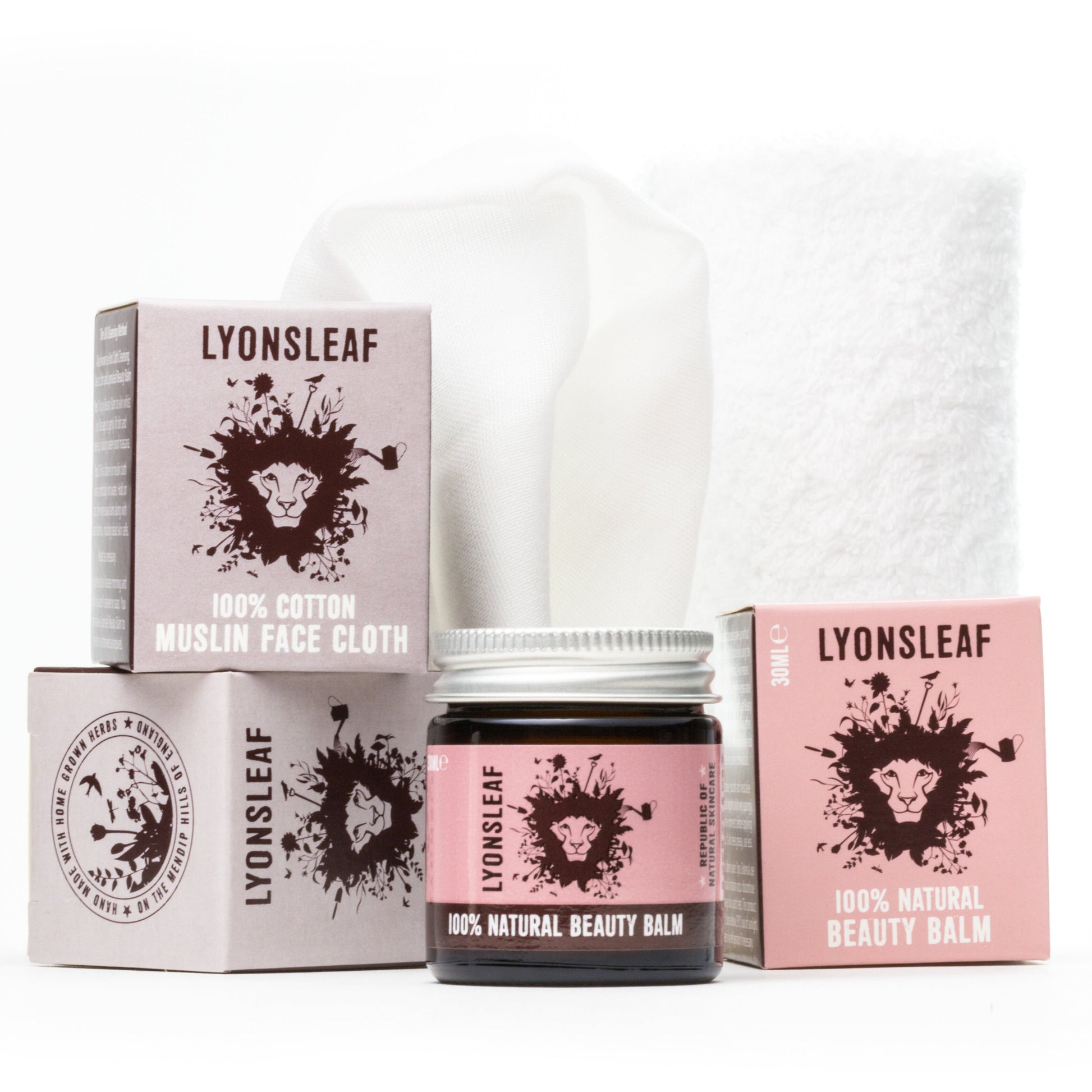 the products that come in the lyonsleaf hot cloth cleansing kit are photographed on a white background. from left to right there is a mauve box that holds the 100% cotton muslin face cloth and is balanced on another purple box. The white 100% cotton cloth is standing upright in a roll in the background. The jar and the box of the 100% natural beauty balm is in the foreground