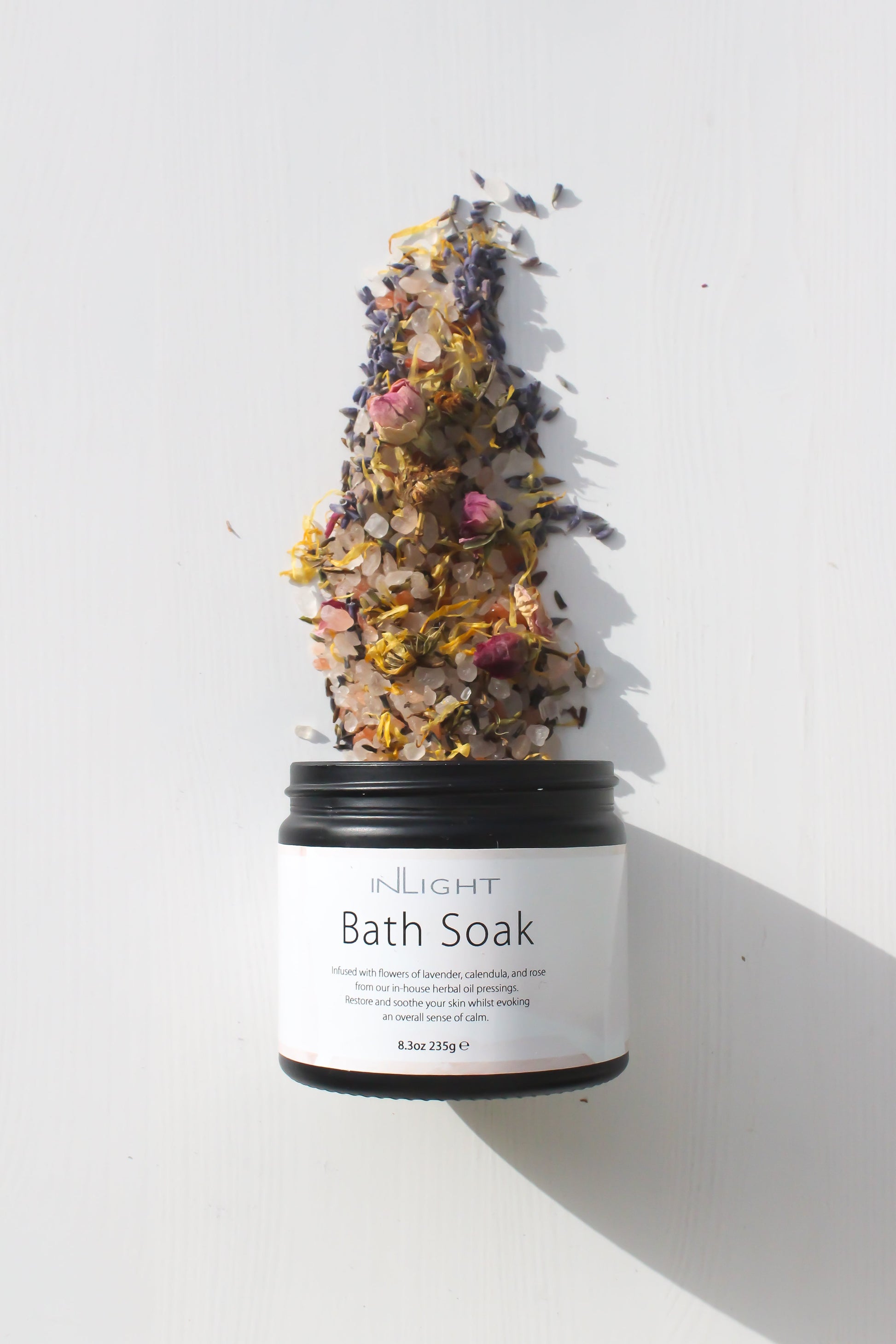 image of inlight beauty bath salts jar laying flat and opened with a spray of the salts and herbs and flowers spilled out of the jar on to the white/grey background