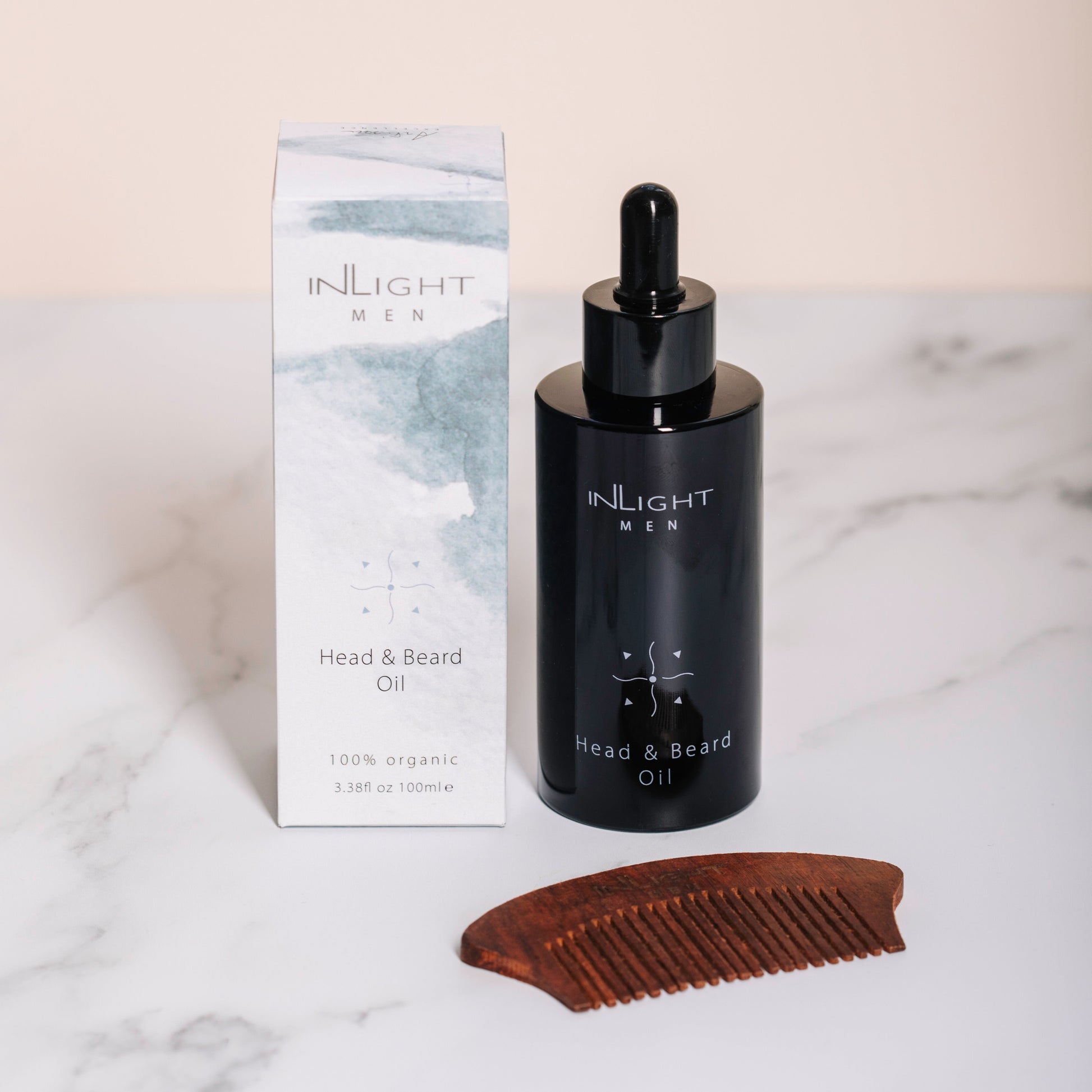 inlight beauty 100% organic head and beard oil in a black recycled miron glass bottle with pipette sitting on a soft grey marble counter with a light pastel pink painted wall behind. Sitting next to the white and watercolour blue grey packaging with a rosewood comb
