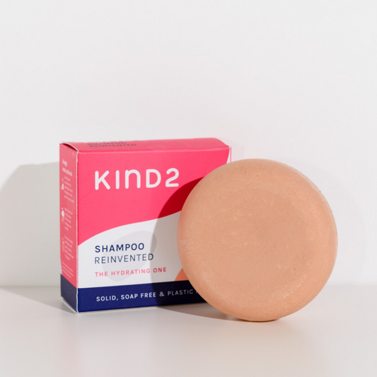 Round pink plastic free KIND2 shampoo bar. Placed in front of its pink, white and navy box. Labelled with Shampoo Reinvented.