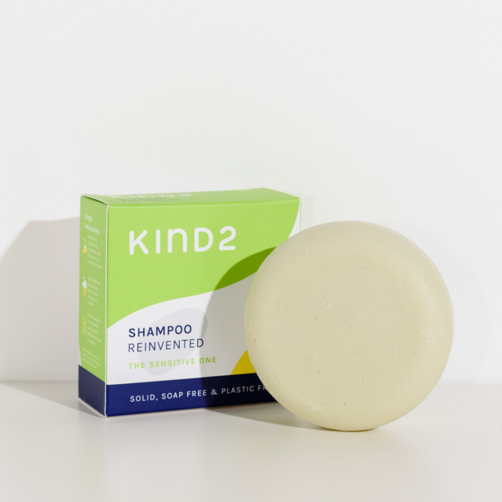 Round off-white plastic free KIND2 shampoo bar. Placed in front of its green, white and navy box. Labelled with Shampoo Reinvented.