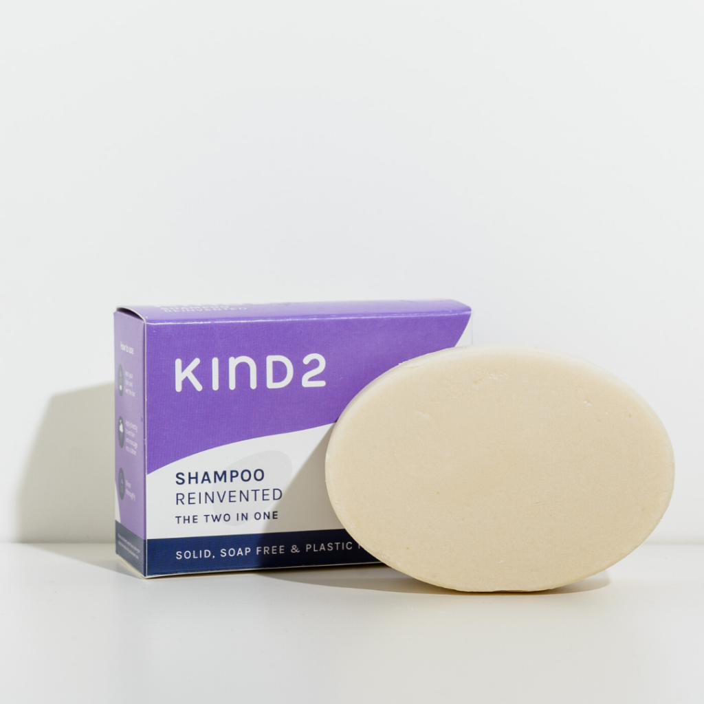 Oval shaped off-white plastic free KIND2 shampoo and conditioner bar. Placed in front of its purple, white and navy box. Labelled with shampoo Reinvented.