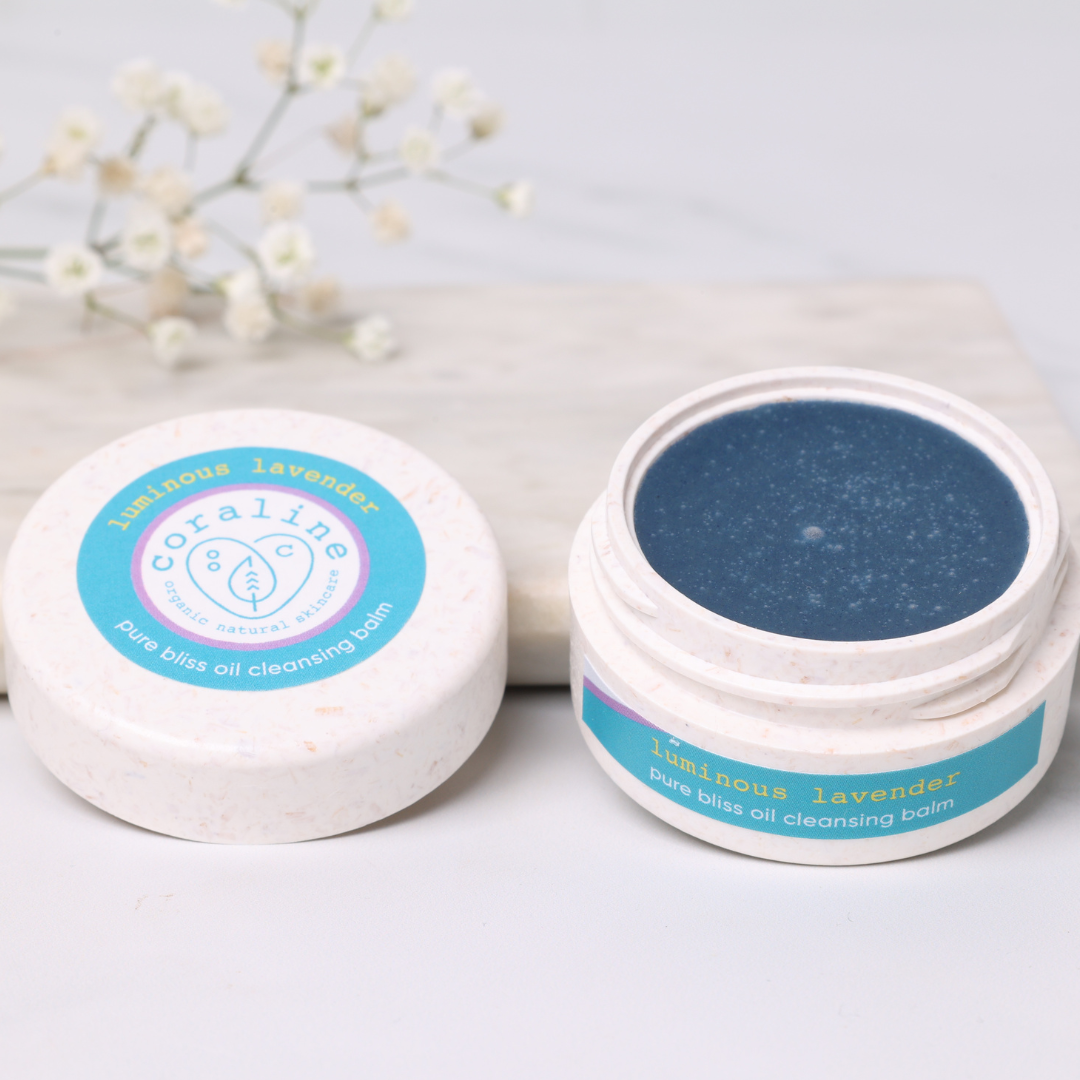 A round jar of Coraline pure bliss oil cleansing balm in luminous lavender is open next to its lid. The product inside is the dark blue balm. The lid and jar label are white with blue accents. In the background are delicate white flowers.