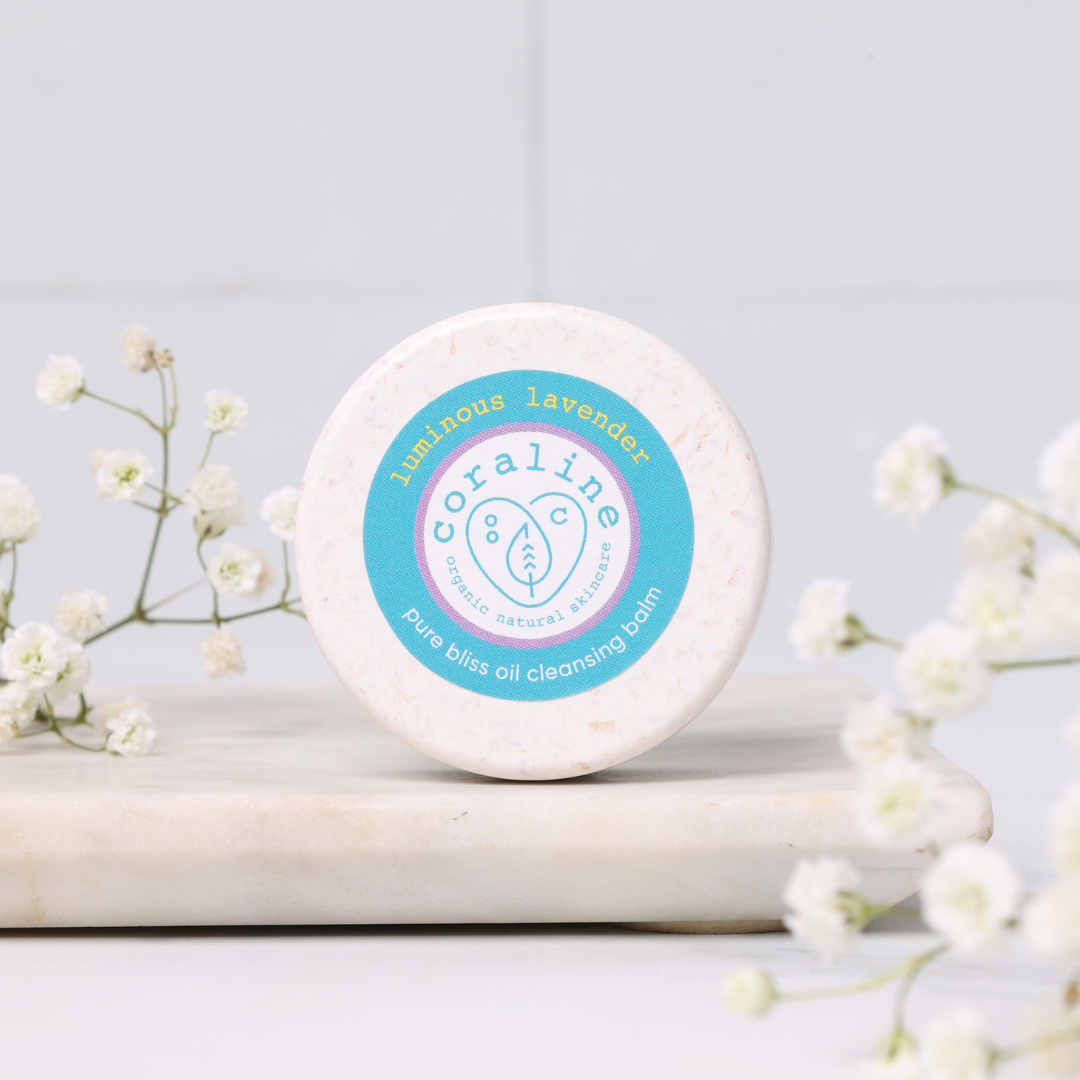 A round container of Coraline Skincare Pure Bliss Oil Cleansing Balm sits on a white marble surface. The label reads "luminous lavender" and is surrounded by small white flowers. The background is softly blurred, emphasizing the product.