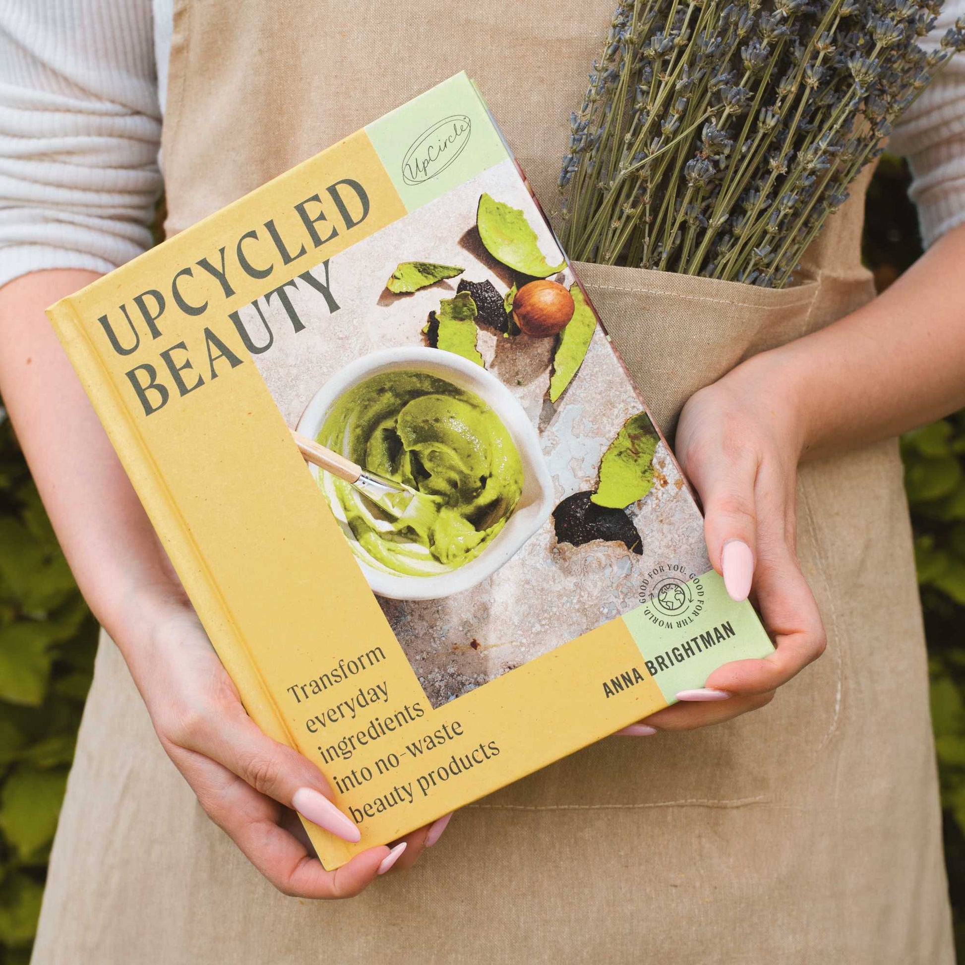 fair skinned person wearing a hessian apron with dried lavender peeking out of the pocket and in her hands she's holding the upcircle upcycled beauty recipe book