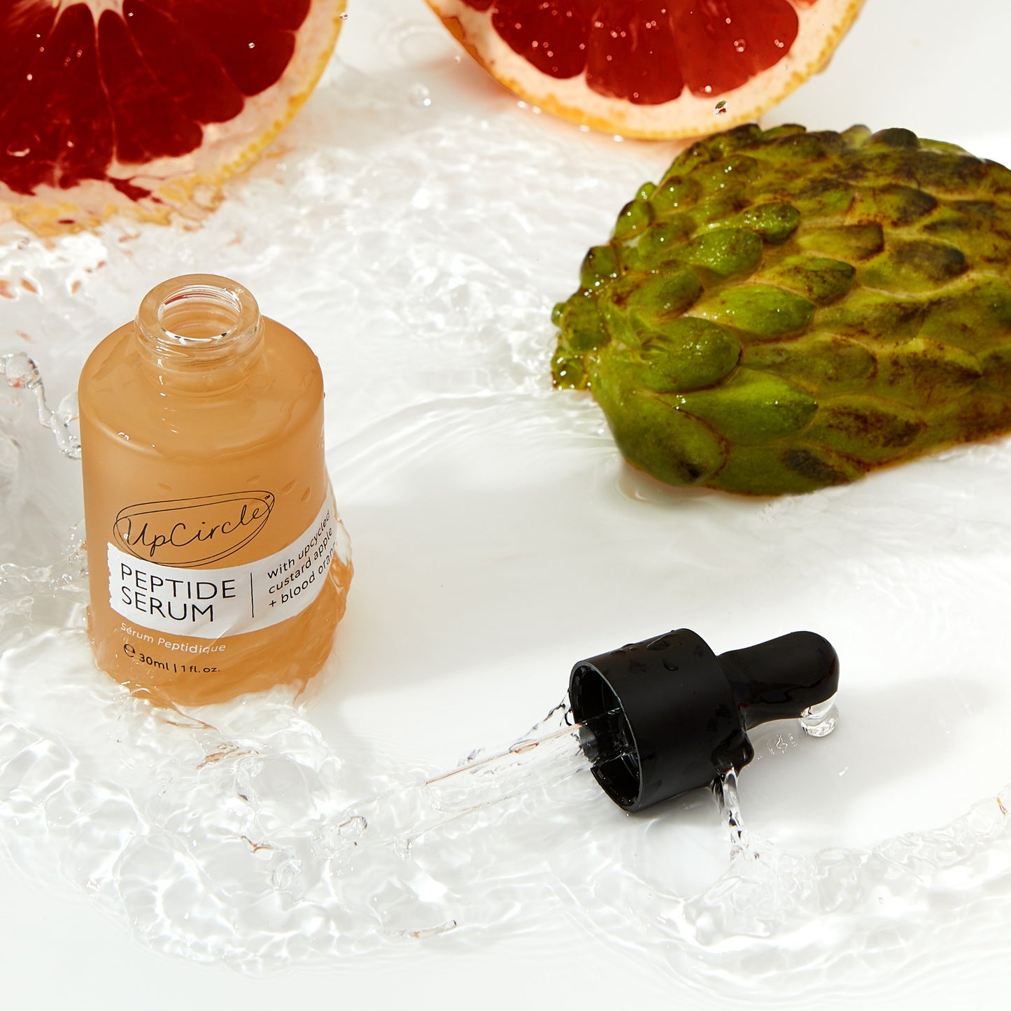 aerial view of upcircle beauty peptide serum bottle with the pipette lid off and placed just in front. both are slathered in the serum and there are custard apples and blood oranges in the background which are the ingredients in the product