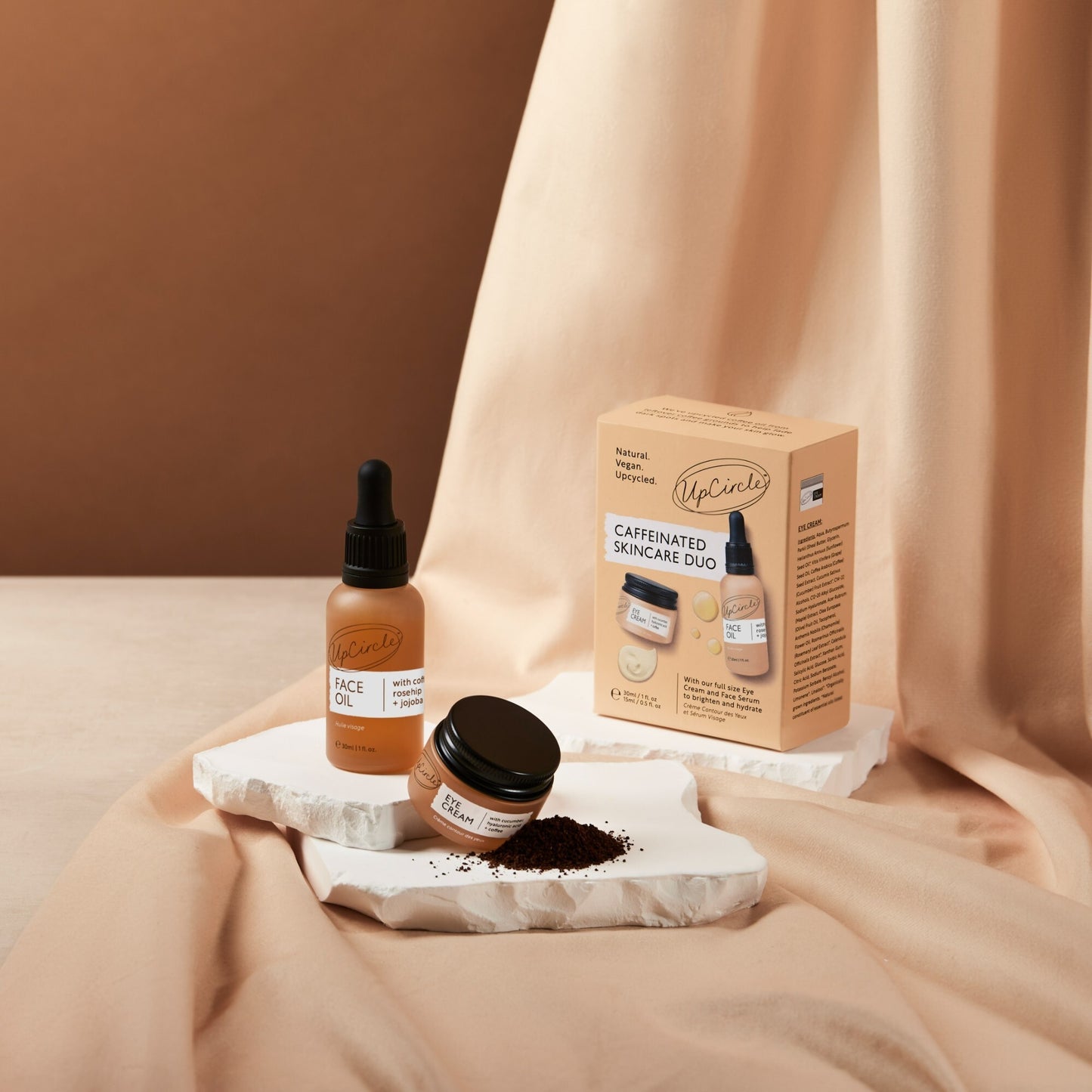 upcircle caffeinated duo products on a peachy brown coloured piece of fabric and a white stone plinth. you can see the box, the face oil bottle and the eye cream jar and there is a pile of coffee grounds to show the ingredients in the products 