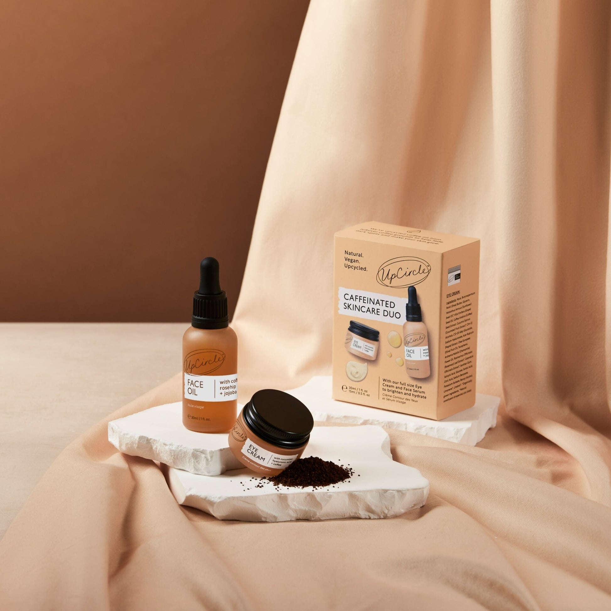upcircle caffeinated duo products on a peachy brown coloured piece of fabric and a white stone plinth. you can see the box, the face oil bottle and the eye cream jar and there is a pile of coffee grounds to show the ingredients in the products 