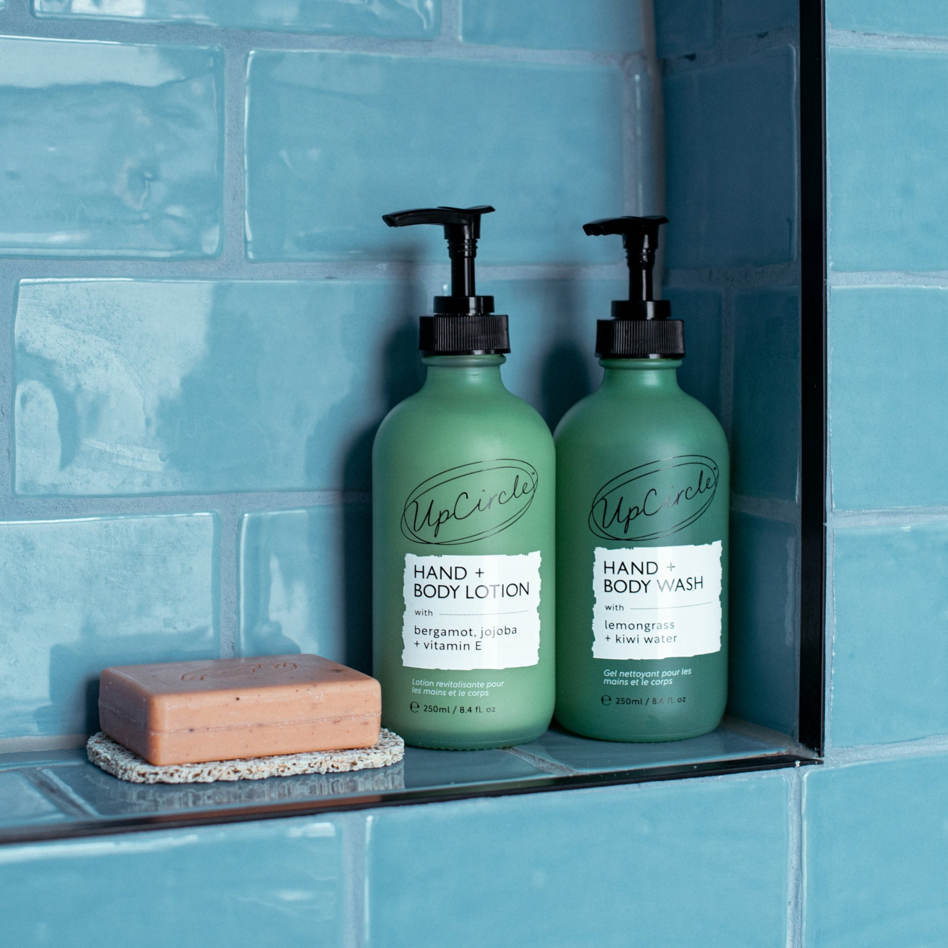 upcircle hand and body lotion and hand and body wash bottles on a bathroom shelf surrounded by tuquoise blue tiles and next to an orange and cinnamon soap bar