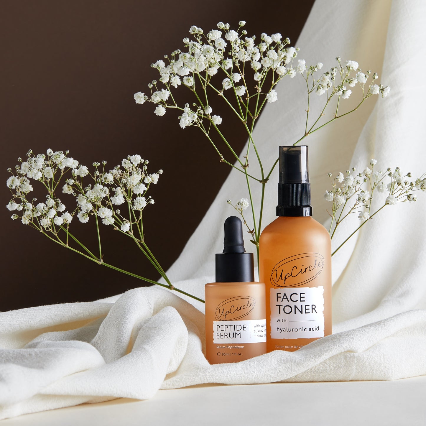 lifestyle image of 2 upcircle products on a white sheet with gypsophelia fronds in the background. features the face toner and peptide serum