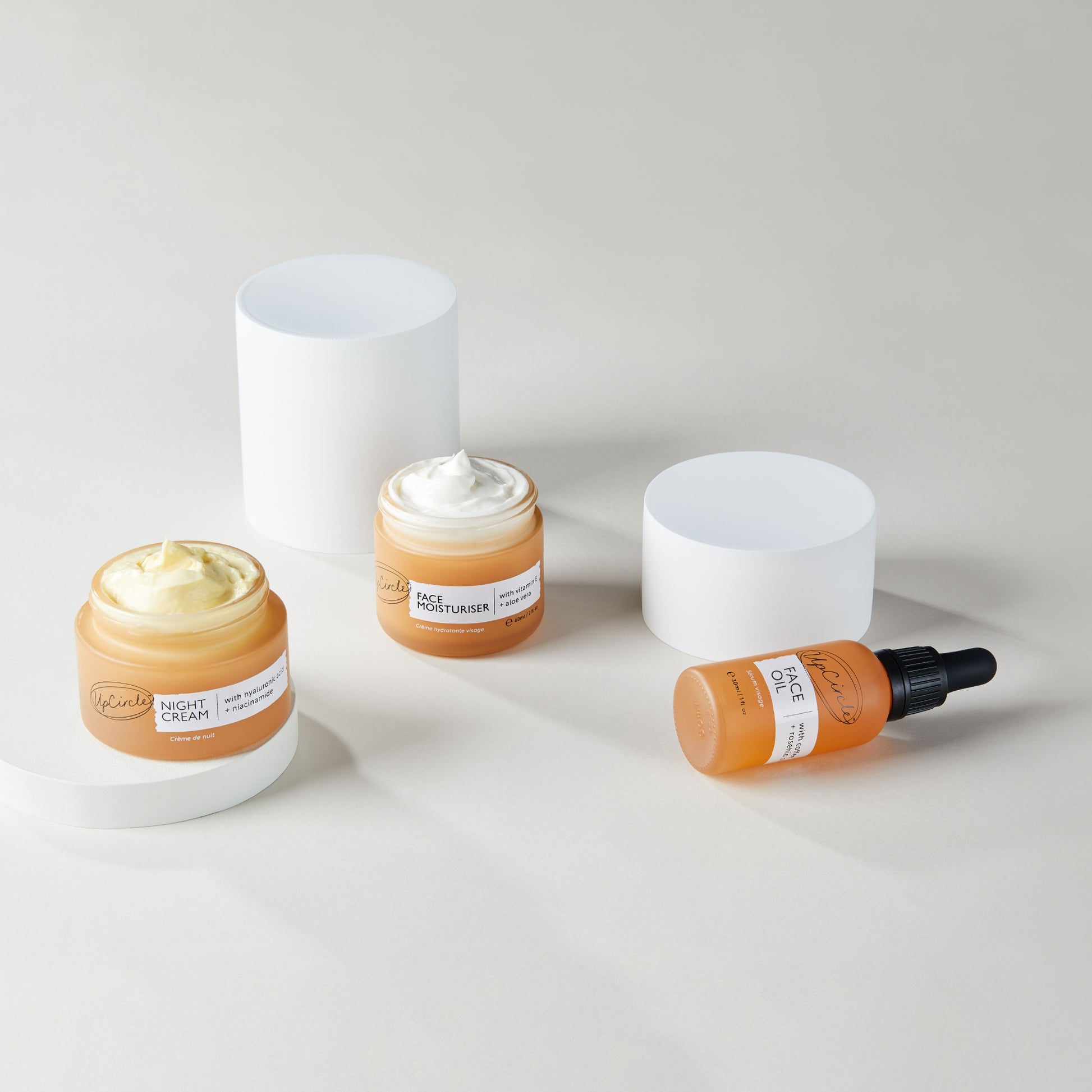 upcircle beauty day and night hydration set photographed on white round plinths. the night cream is on the lowest plinth, the moisturiser is in front of the highest plinth and the face oil is on its side in front of the medium sized plinth