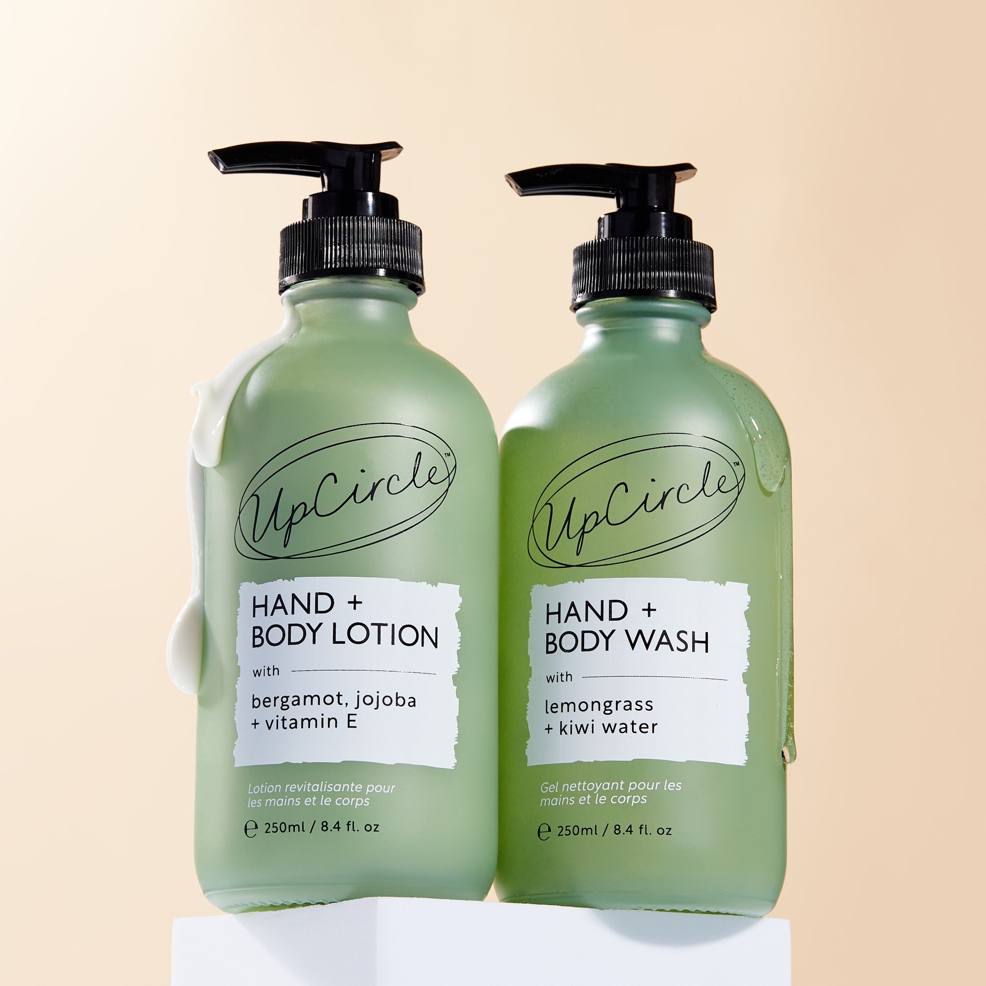 green frosted glass bottles of upcircle hand wash and hand and body lotion on a white plinth with a peachy background