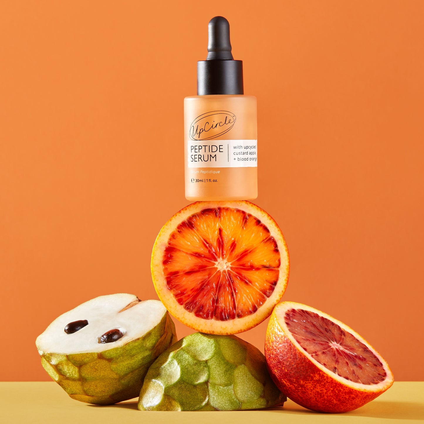 upcircle peptide serum balancing on sliced of fruit custard apples, and blood oranges with an orange background