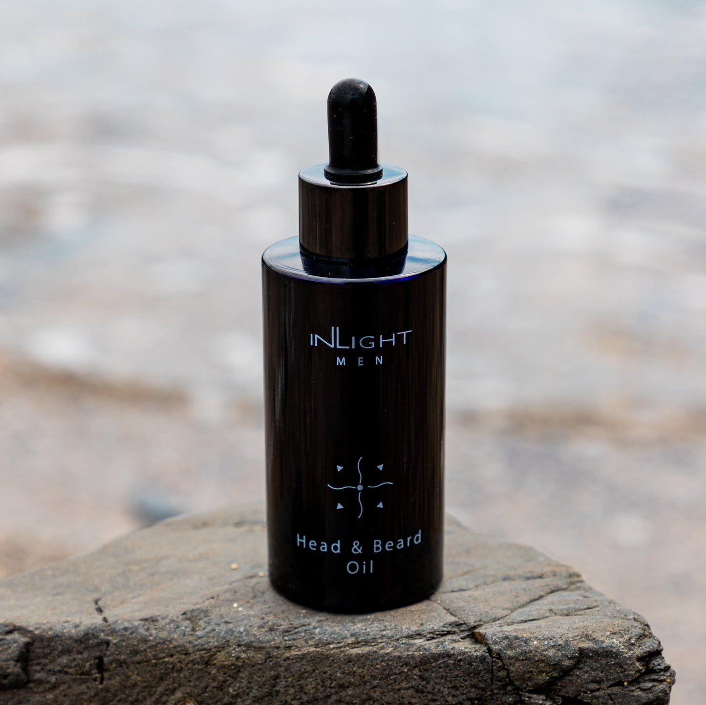 inlight beauty 100% organic head and beard oil in a black recycled miron glass bottle with pipette sitting on a grey rock with a blurred beach setting in the background