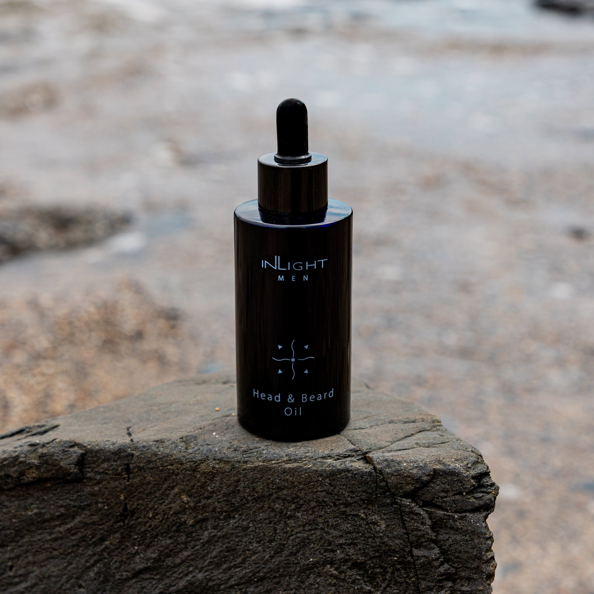 inlight beauty 100% organic head and beard oil in a black recycled miron glass bottle with pipette sitting on a grey rock with a blurred beach setting in the background where you can see pebbles and placid water