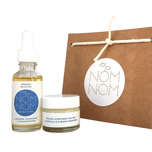 photo of nom nom skincare mini gift set for face and body on a white background. featuring left to right; a small bottle of organic relax oil with lavender, chamomile and frankincense in a clear glass bottle with a white pipette, an organic everybody butter with calendula and evening primrose in a glass jar with white screw cap lid and a brown kraft paper gift bag with the nom nom logo on it