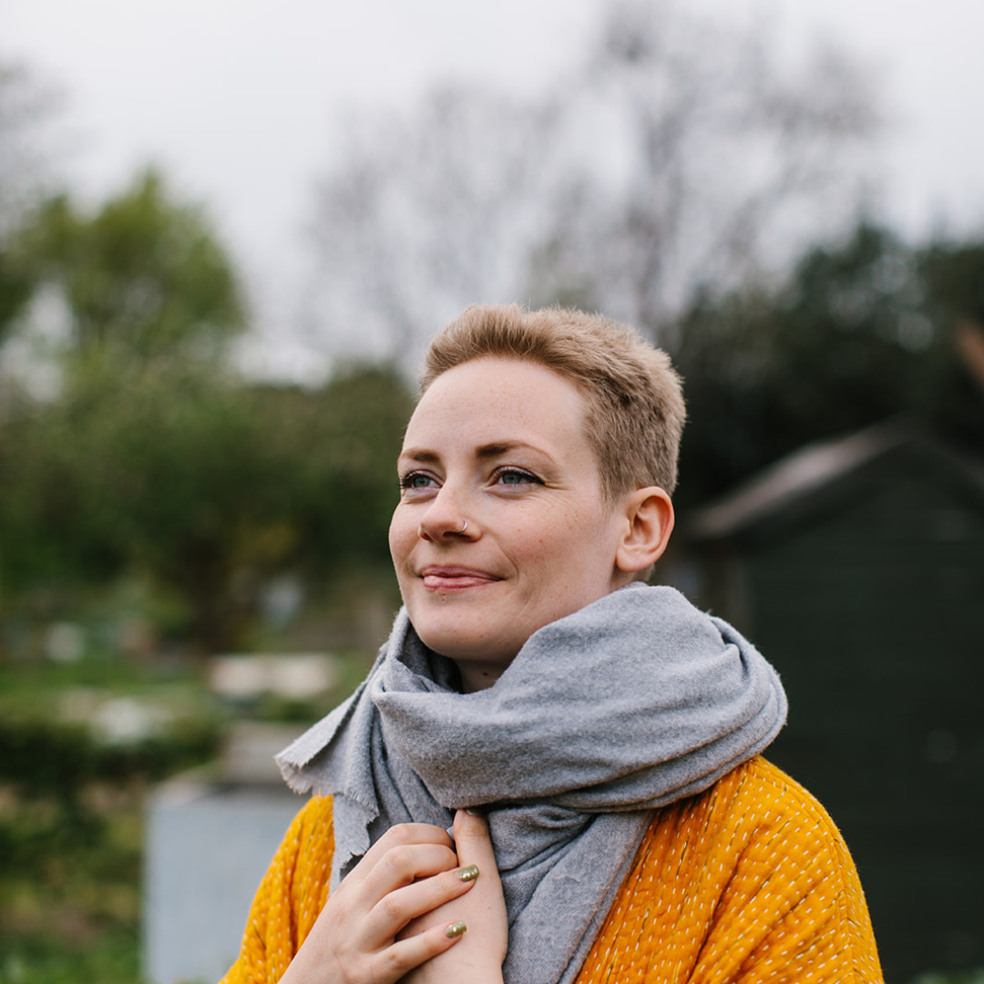 natasha richardson is a medical herbalist and founder of hormonal support products range forage botanicals. she is photographed amongst green shrubbery with white blossoms on it wearing an orange yellow jacket and grey scarf. she has short strawberry blonde hair, blue eyes and pale skin