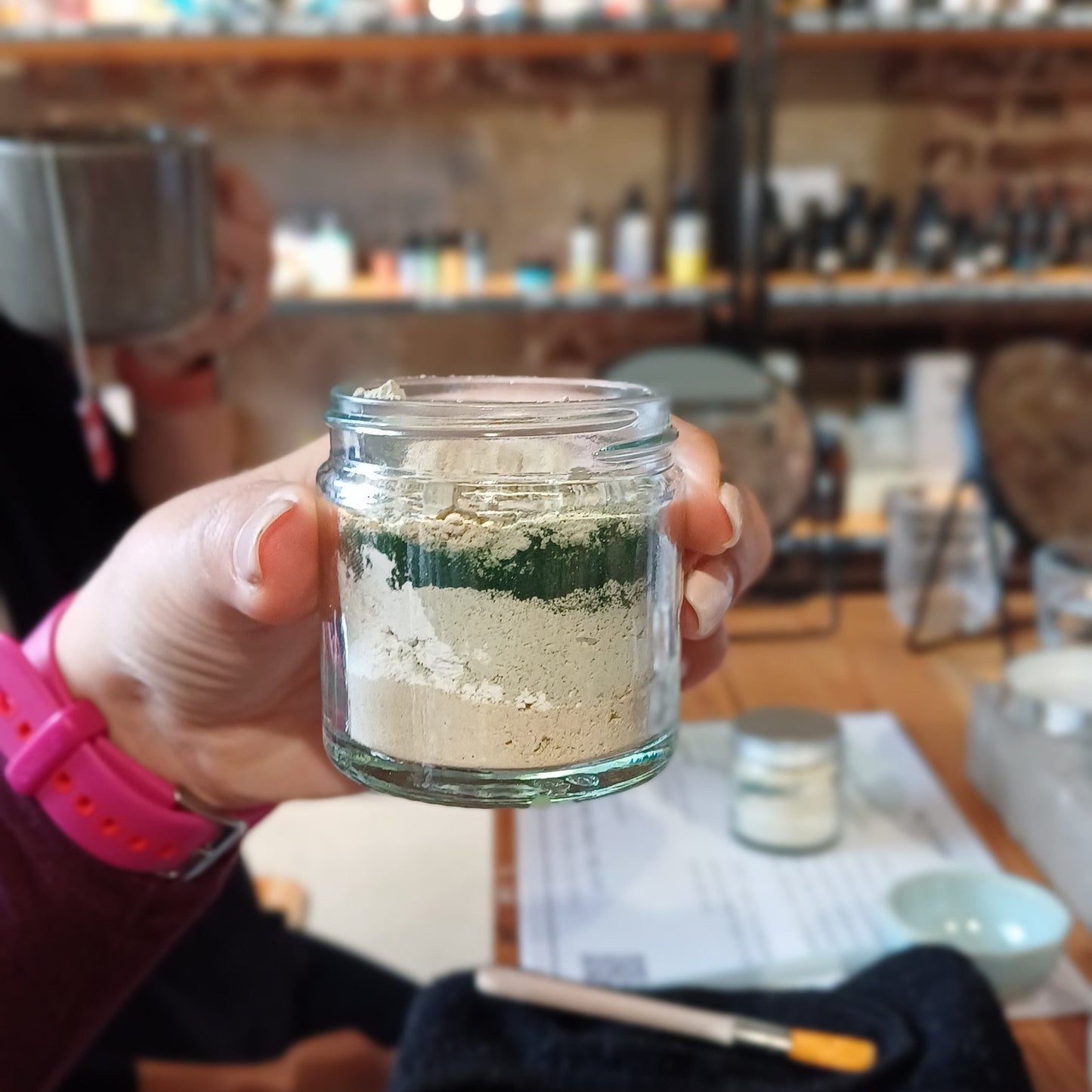 woman with pink rubber watch strap and light pink nail varnish is holding up the clay face mask before it is blended. the clay face mask is in a clear glass jar and shows the layers of different clays and active skincare ingredients used to craft your own clay face mask in our make your own skincare workshop