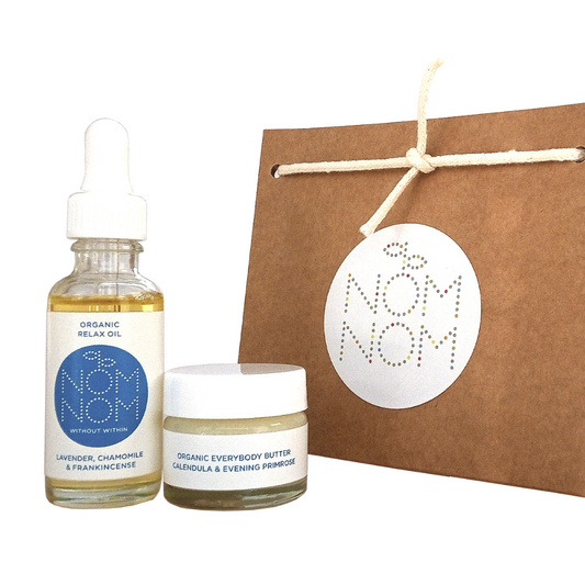 photo of nom nom skincare mini gift set for face and body on a white background. featuring left to right; a small bottle of organic relax oil with lavender, chamomile and frankincense in a clear glass bottle with a white pipette, an organic everybody butter with calendula and evening primrose in a glass jar with white screw cap lid and a brown kraft paper gift bag with the nom nom logo on it