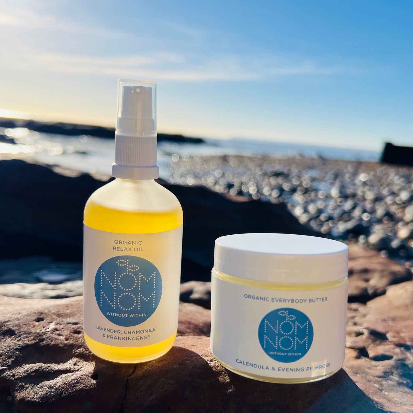 photo of nom nom skincare organic body oil and body butter sitting on the edge of a rock pool. the background is blurred but you can make out a pebble beach and the sea in the background as the products sit in the evening sun