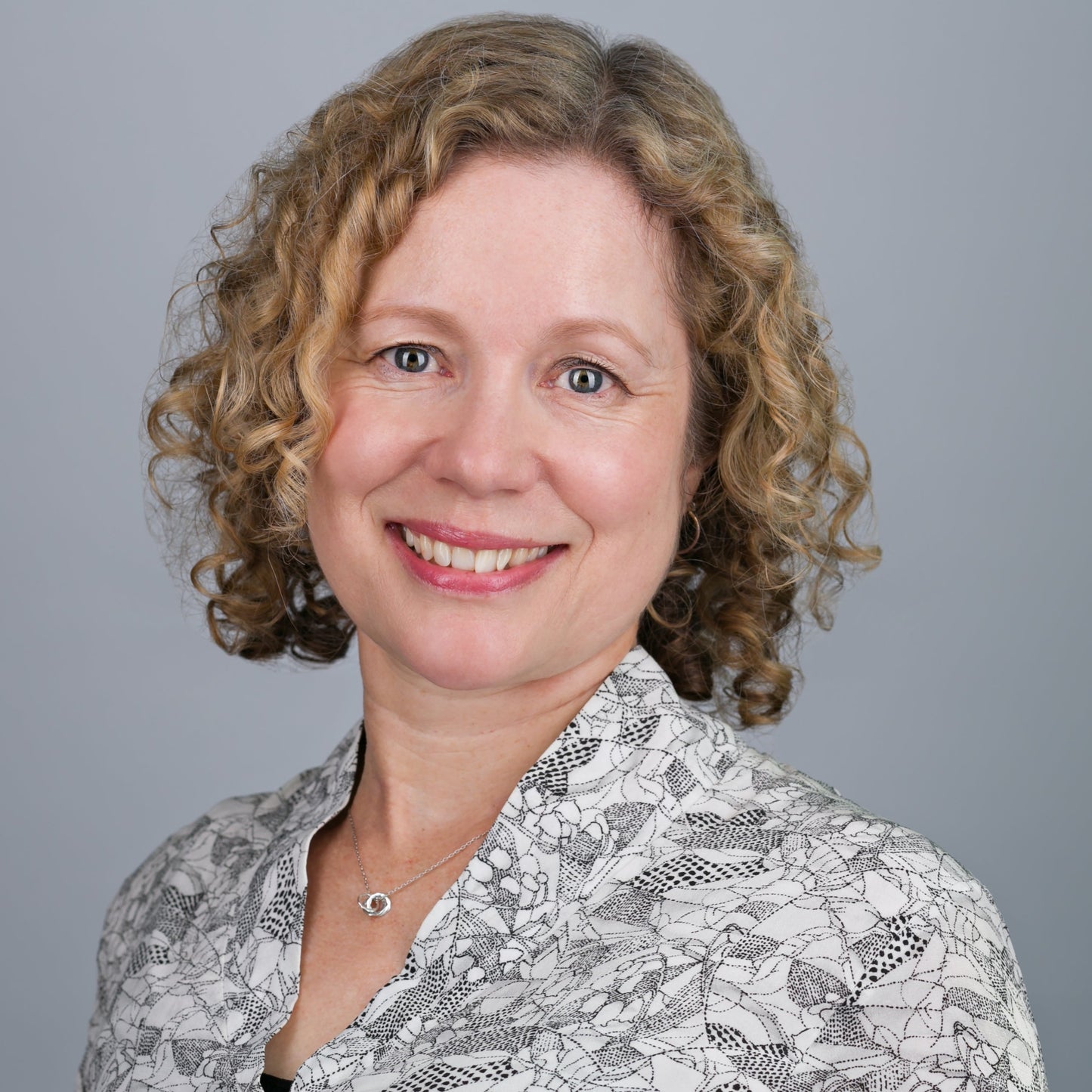 Photo of Jayne Russell maker of Nom Nom Skincare products for babies, pregnancy and everybody. Jayne has a curly blonde bob and it wearing a black and white graphic printed blouse set against a warm grey background