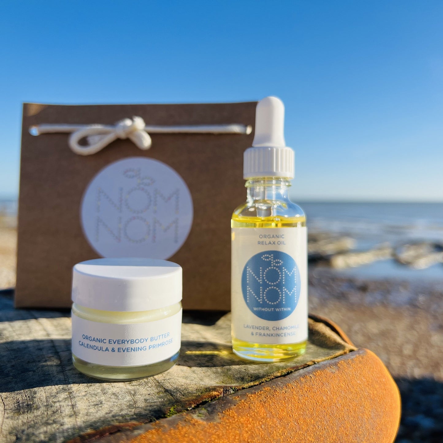 the nom nom skincare face and body relaxing skincare gift set is sitting on a wooden and metal surface that has rusted. in the foreground you can see the glass pipette bottle and jar containing the gentle skincare and the brown kraft gift bag behind it. in the background, although it is blurred, you can see that the photo is taking place on a sunny pebble beach with the tide out and pools of water everywhere