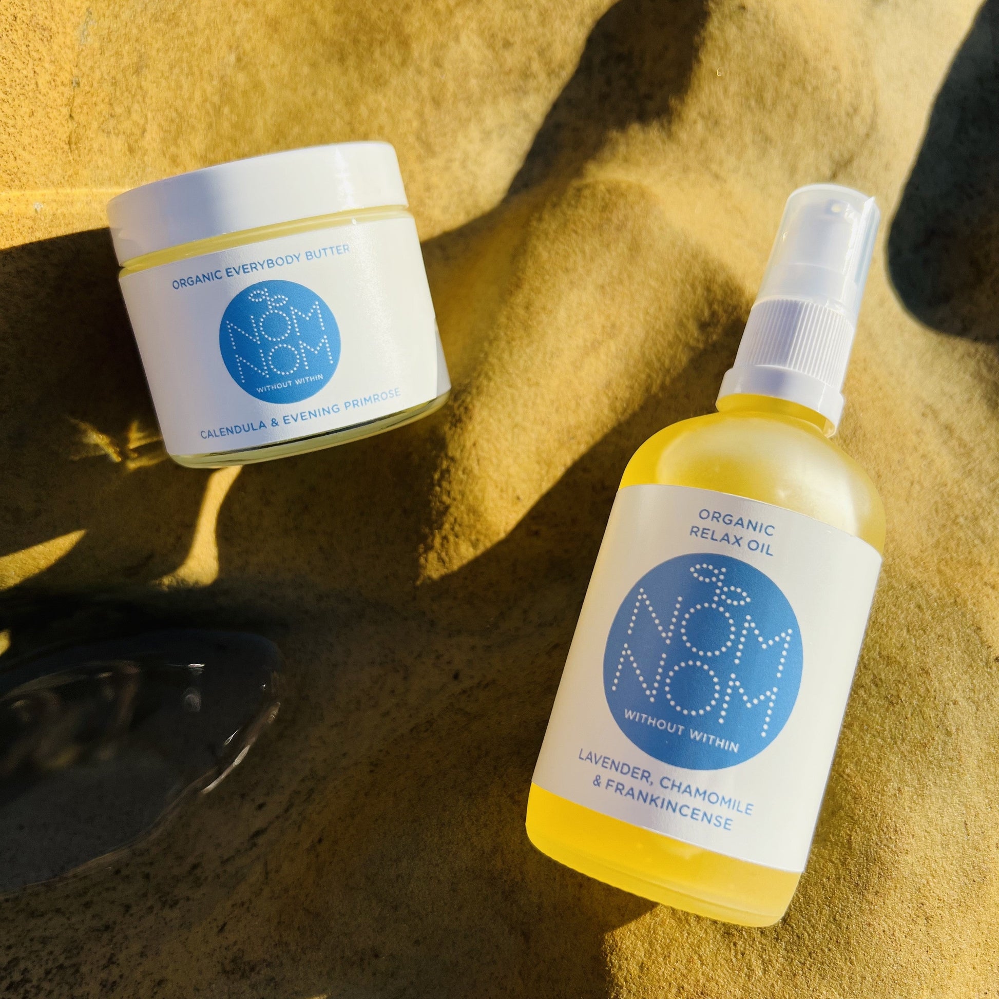 lifestyle photo of nom nom skincare's everybody body butter and body oil placed on a bed of sand on the beach and in warm sunlight