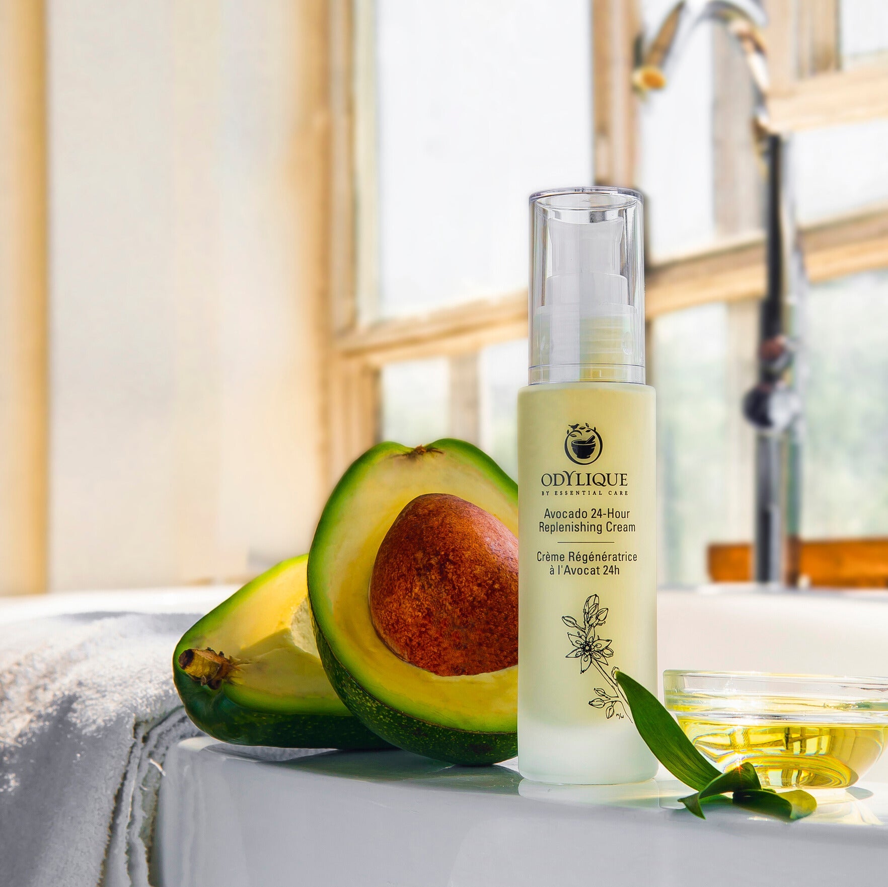 face cream made with avocadoes sitting on the side of a white sink with a chrome tap in the background. the organic moisturiser is next to a small bowl of avocado oil and two slices of avocado with the stone in the middle of one of them