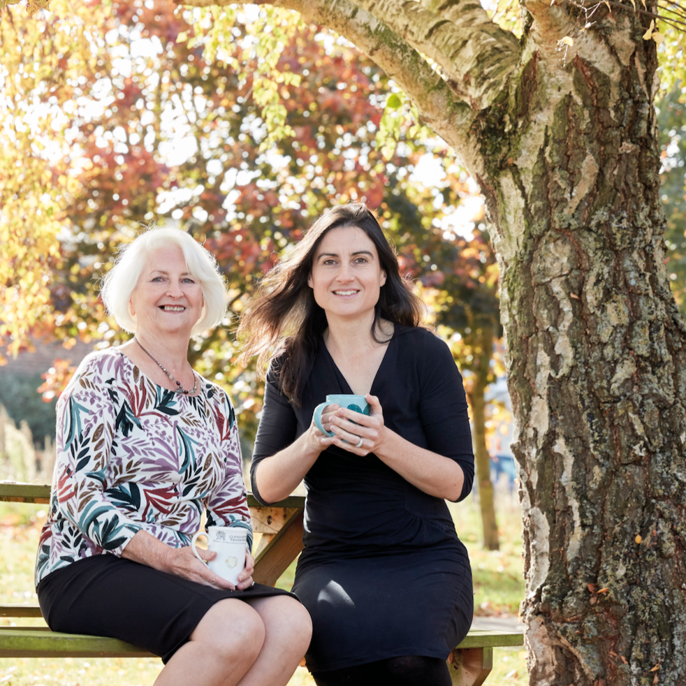 Portrait of Margaret Weeds and her daughter Abi, sitting on a bench outdoors next to a tree. Both are holding mugs with both hands, enjoying a moment of relaxation and warmth