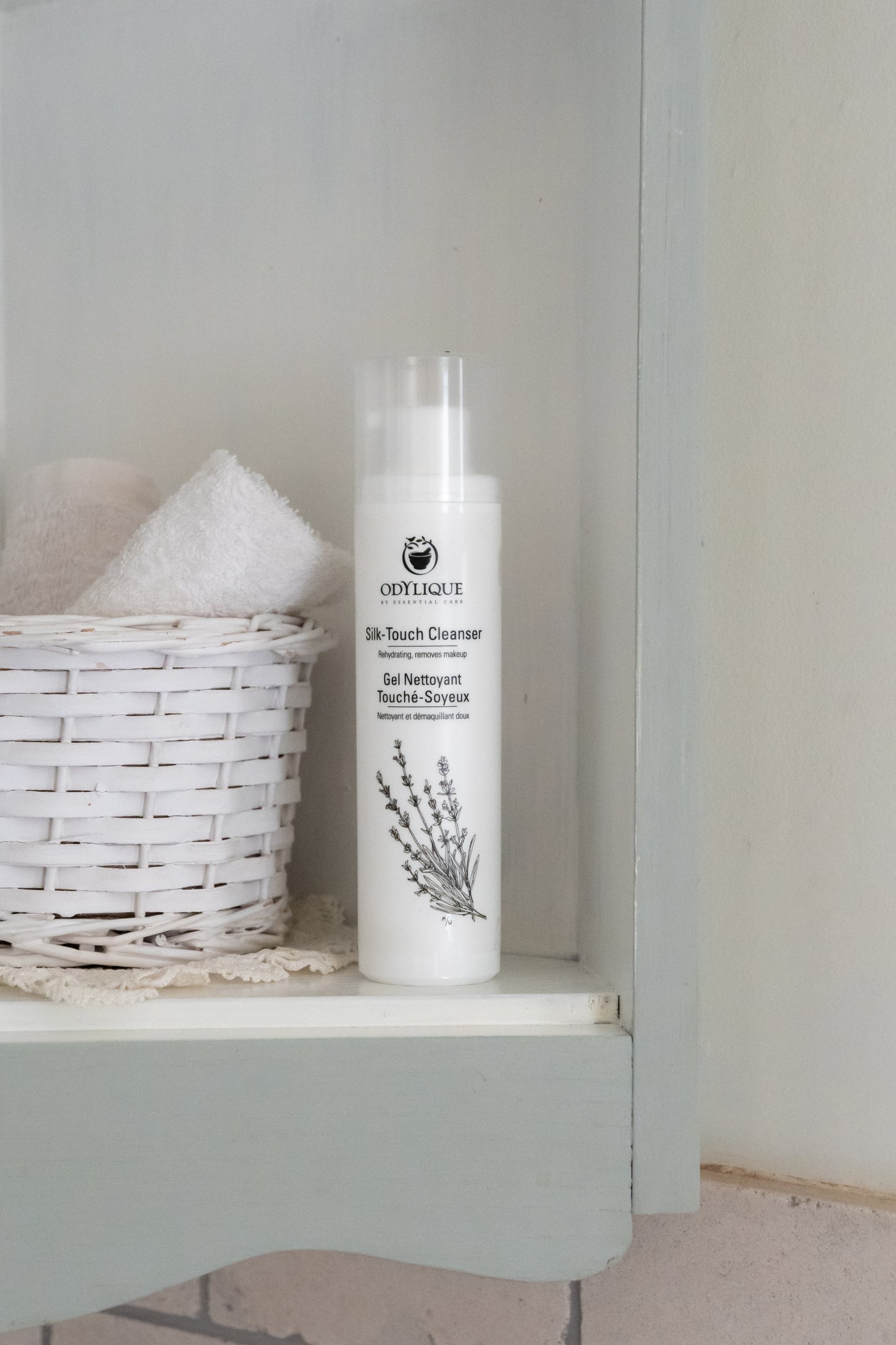 odylique silk touch cleanser for very dry, mature skin sitting on shelf in bathroom next to a white wicker basket with white fluffy face towels peeking out