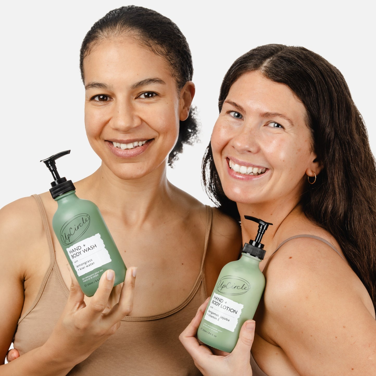 2 women with brown hair and flesh coloured vest tops holding upcircle's green glass bottles with hand and body lotion and wash in them