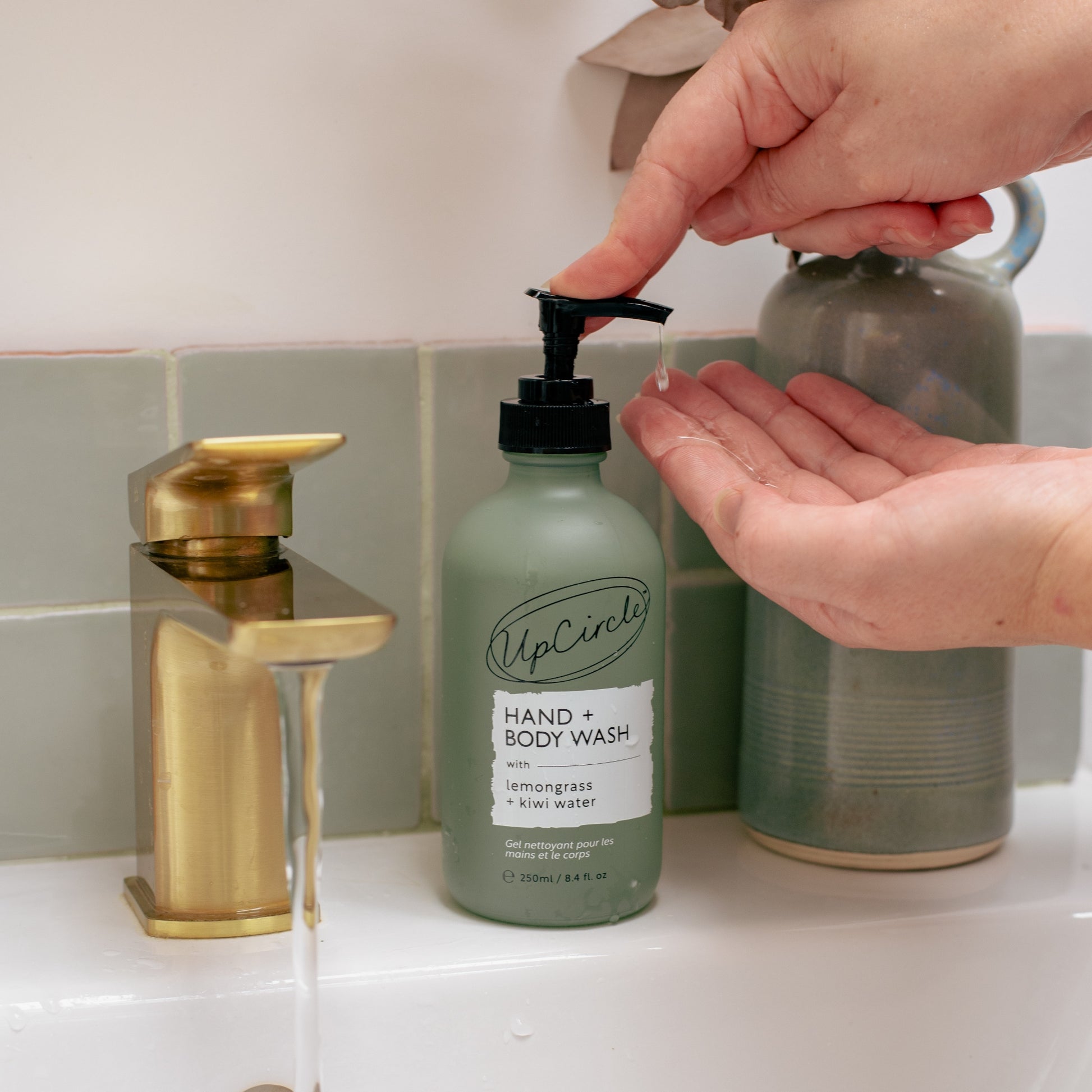 hand and body wash green glass bottle on a bathroom sink with gold tap with running water next to it. hands are dispensing the soap as if they are going to wash their hands