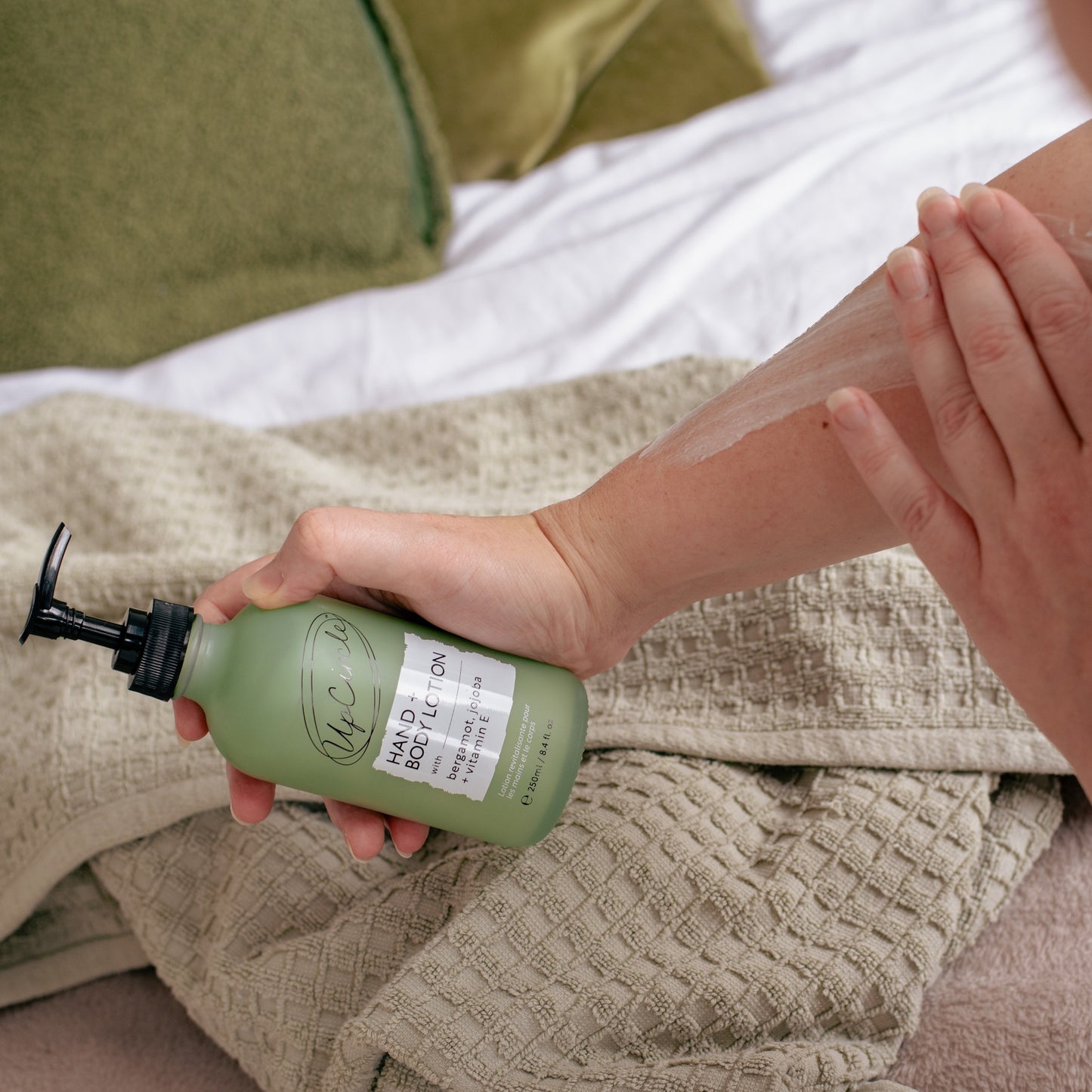 woman applying upcircle hand and body lotion to their arm in the background are green cushions on a bedspread and taupe coloured towel on the bed as if they have just got out of the shower