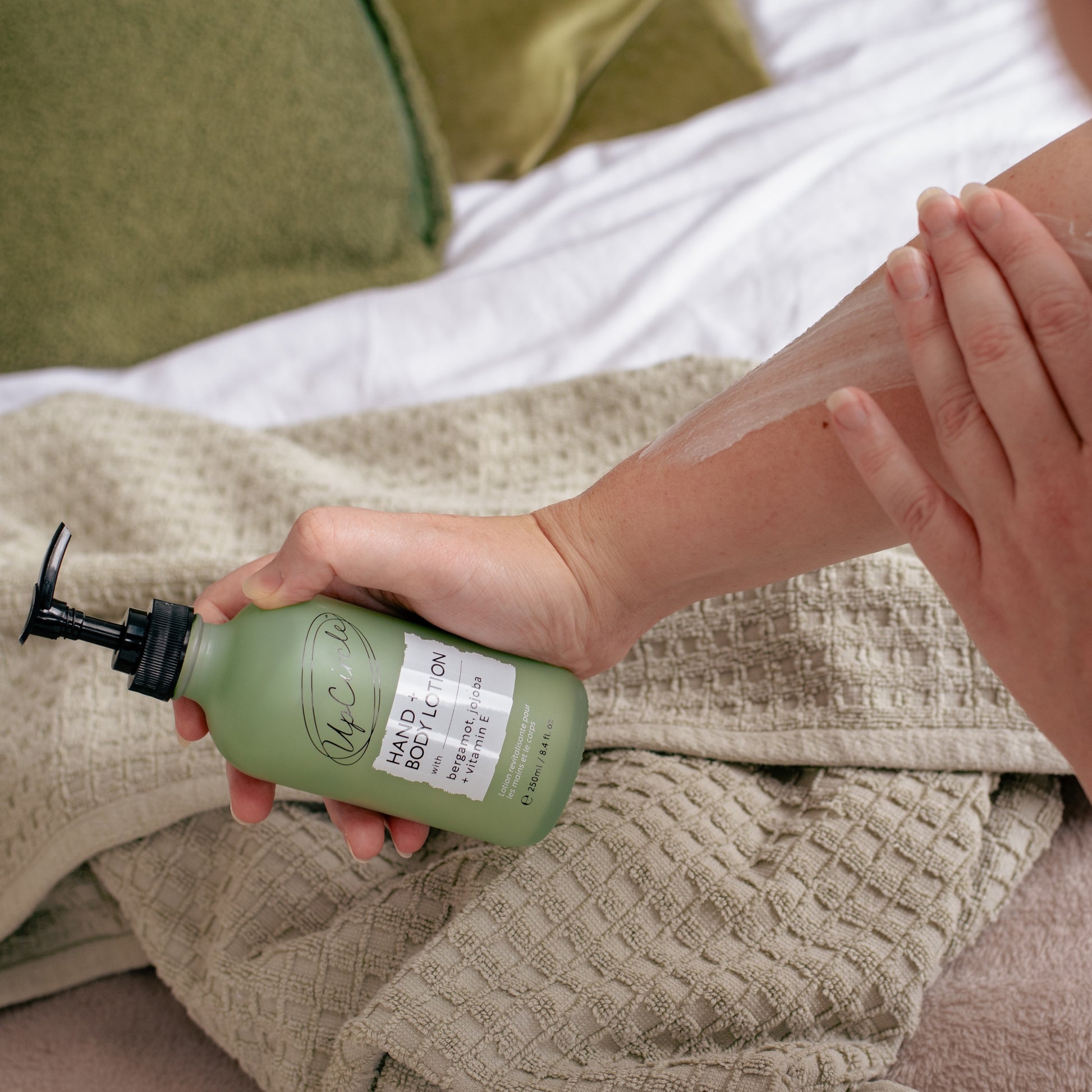 woman applying upcircle hand and body lotion to their arm in the background are green cushions on a bedspread and taupe coloured towel on the bed as if they have just got out of the shower