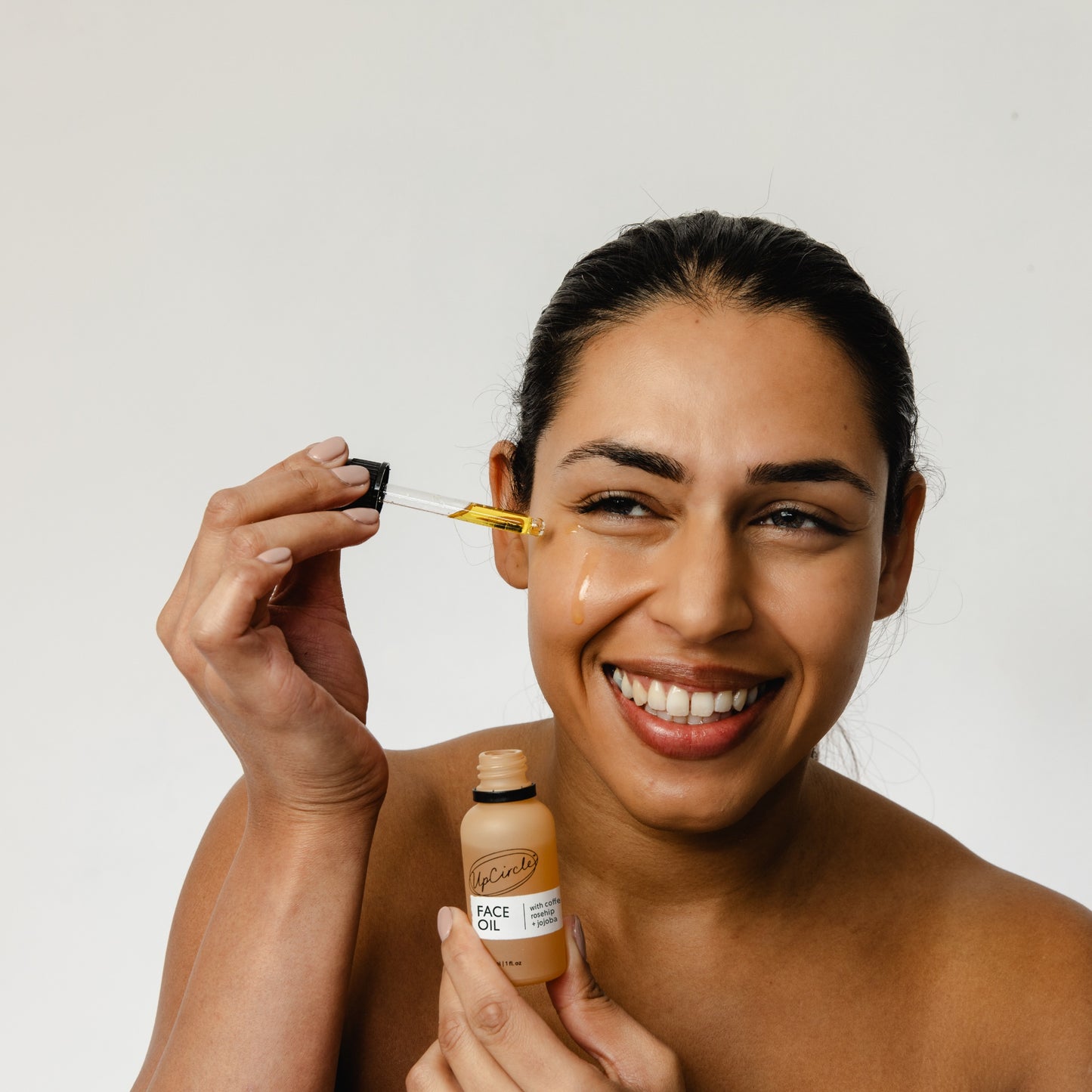 woman with brown skin and brown hair applying the upcircle beauty face oil to her face with the pipette. the oil is dripping down her face 