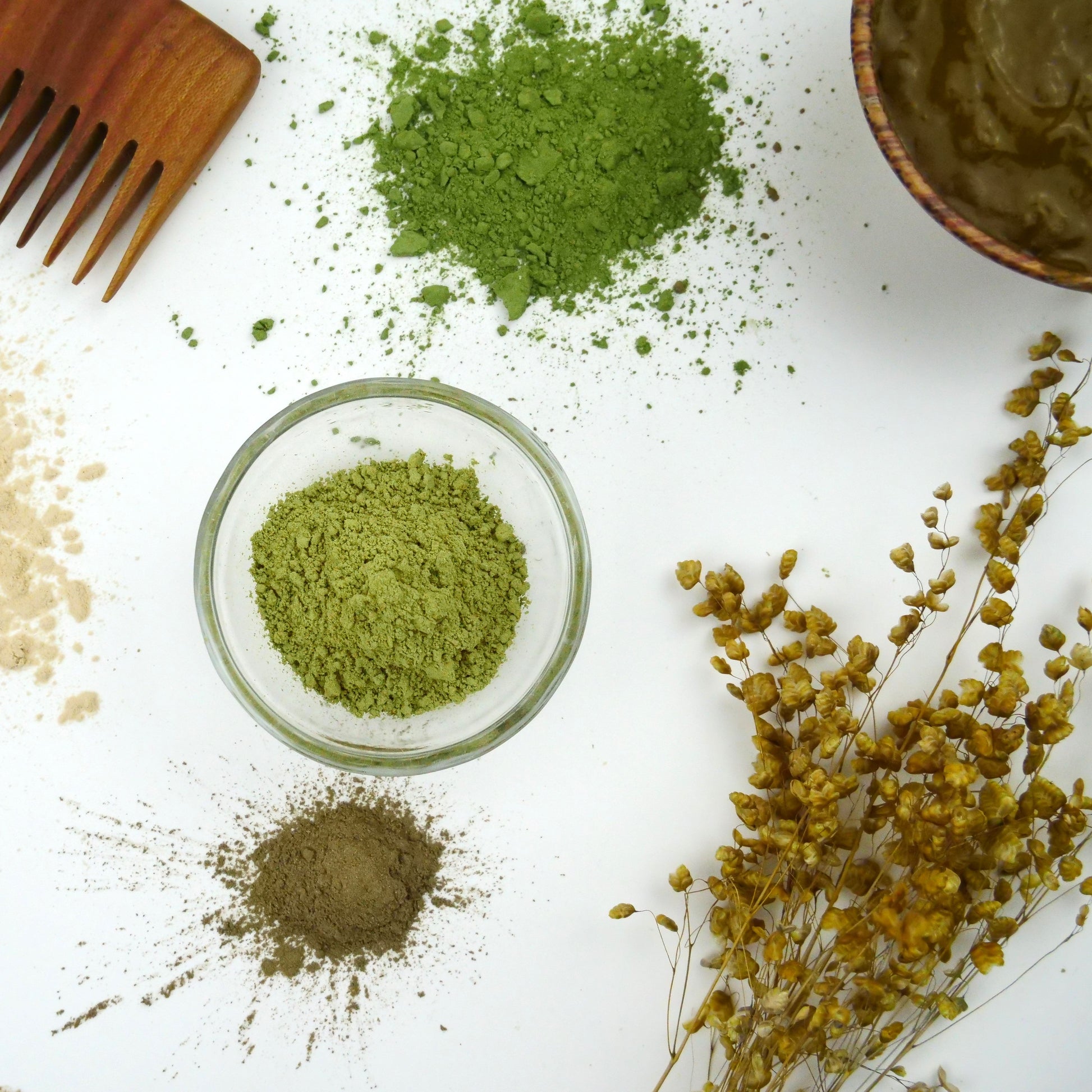 flat lay of ingredients used in it's pure natural henna hair dye. on a white background you can see bright green powders on the table and in a glass bowl, a wooden comb and a dried flower frond. in the corner in a wooden bowl you can see the dye has been mixed with water to create a paste to apply to the hair