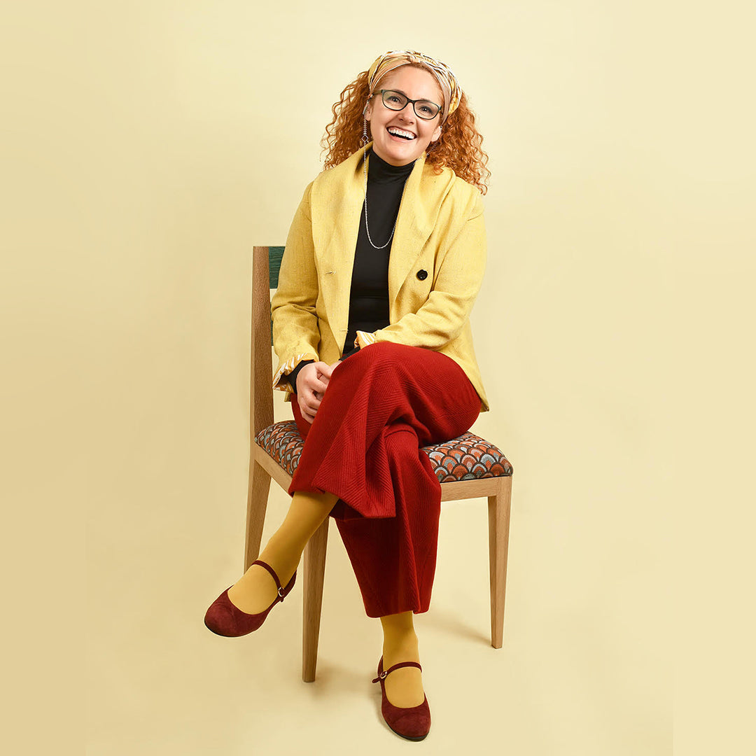 Whitfords skincare founder Paula ortega sitting on chair wearing yellow and red outfit. red mary jane shoes, yellow socks, red wide legged trousers, black polo neck and yellow blazer. she has curly orange hair tied back in a multi coloured head scarf and is wearing black glasses