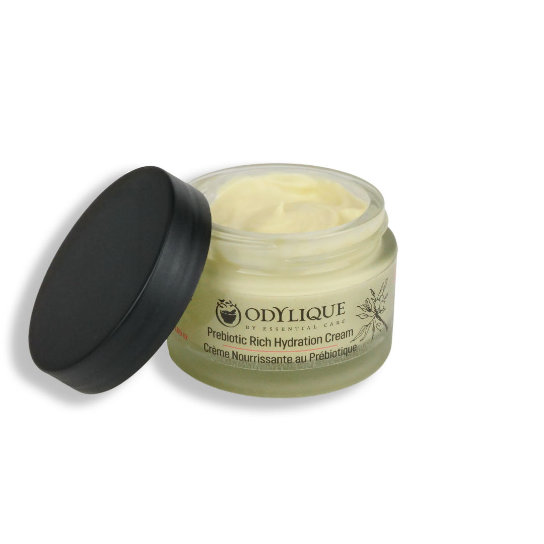 An image of the “Odylique Prebiotic Rich Hydration Cream” against a white. The glass jar partially open with the lid resting against it, showing the creamy texture of the product.