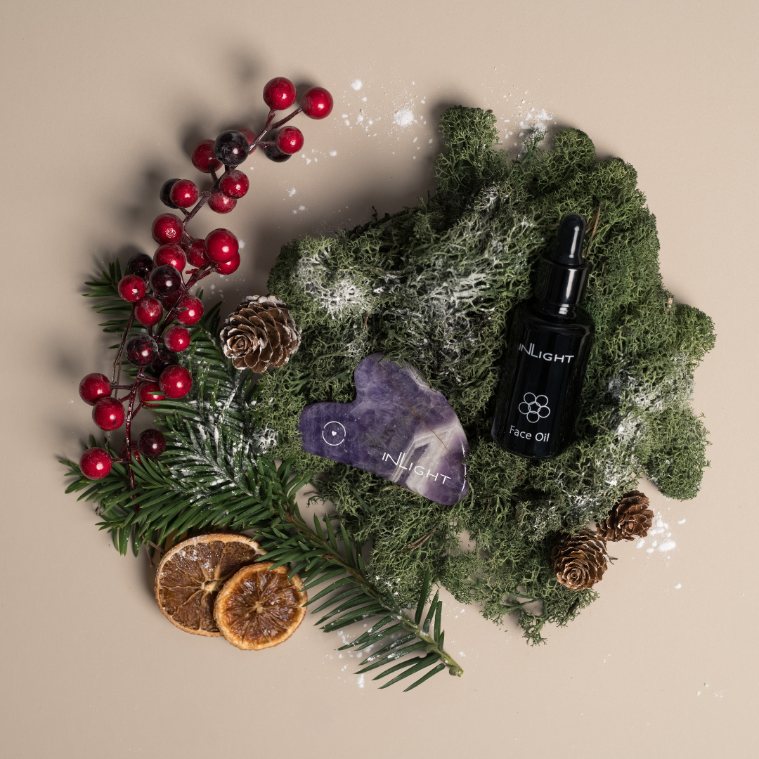inlight beauty's black pipette bottle containing facial oil sitting next to a purple amethyst gua sha tool. both products are sitting on top of dried sea moss with pine cones, pine banches, red berries and dried orange slices
