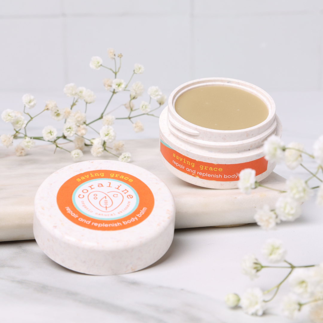 An open pot of "Saving Grace" body balm from Coraline Skincare is placed on a marble surface with white baby’s breath flowers scattered around. The white container has an orange and white label and the balm inside is light yellow.