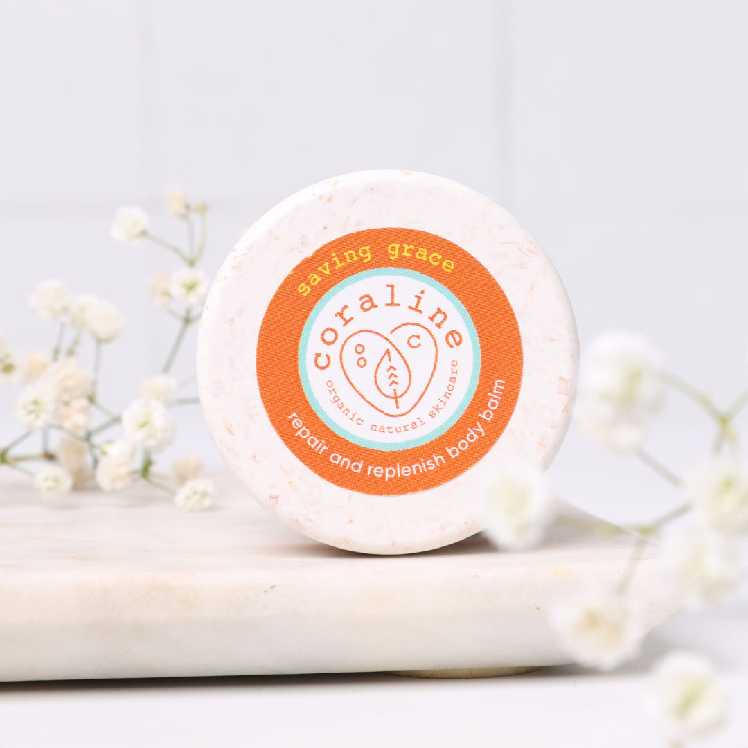 A round container of Coraline organic natural skincare's "Saving Grace" repair and replenish body balm is displayed on a marble surface. The Biodegradable pot has an orange label. White flowers are arranged around the container.