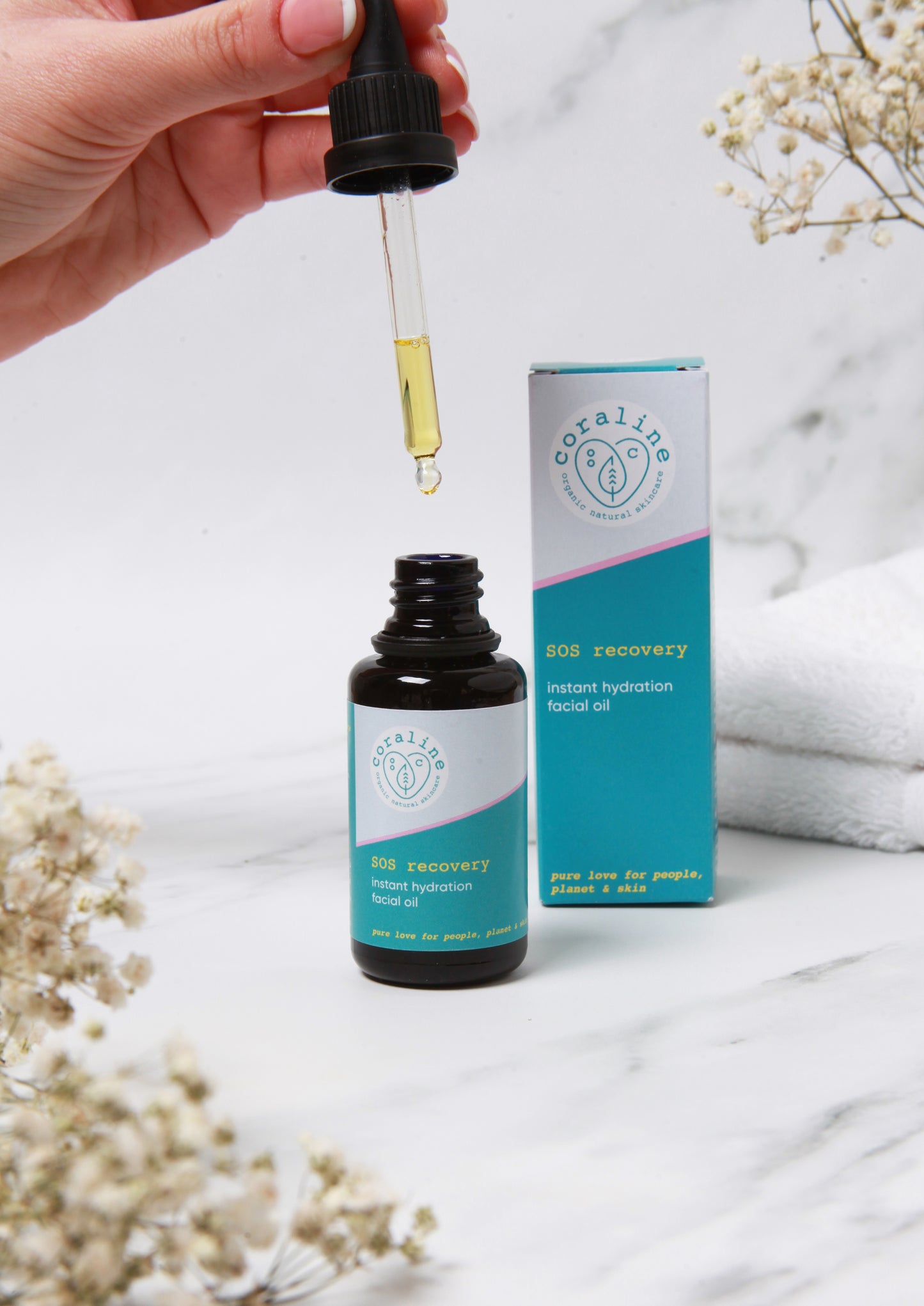 A hand holds a dropper above a small bottle of Coraline Skincare “SOS recovery instant hydration facial oil”. The bottle is open, and the dropper is filled with yellow oil. The product's packaging is visible in the background against a marble surface with white flowers nearby.