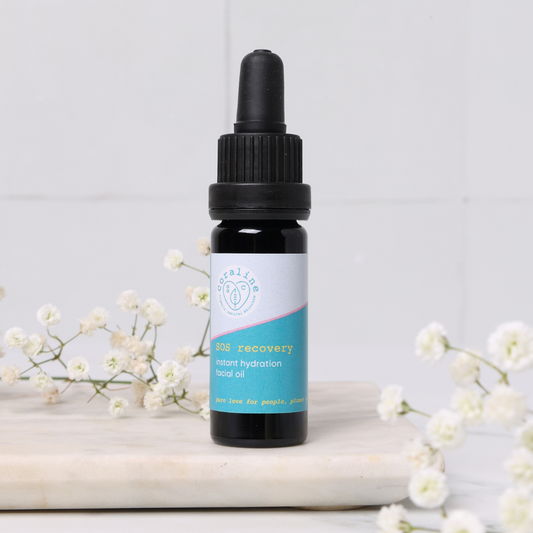  A small dark glass bottle of SOS recovery oil with a light blue label. The bottle sits on a marble surface with baby's breath flowers around it