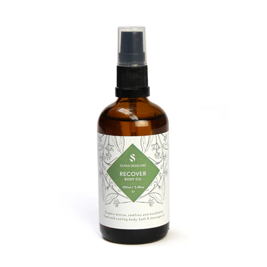 Silvan Skincare Recover Body Oil in an amber glass bottle with a white label and a green diamond in the centre with white text listing the product and brand. The label also has  black botanical illustrations.