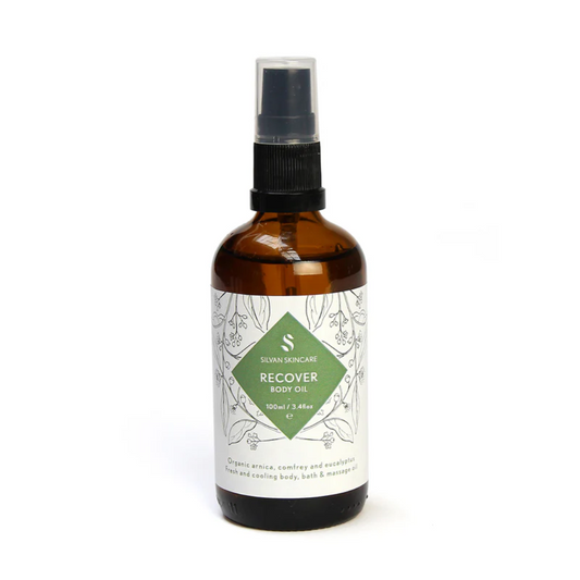 Silvan Skincare Recover Body Oil in an amber glass bottle with a white label and a green diamond in the centre with white text listing the product and brand. The label also has  black botanical illustrations. 