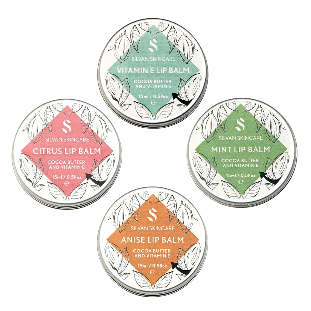 Four tins of Silvan Skincare lip balm are displayed in a square formation. Each tin has a different label (Top-Bottom-Clockwise): Vitamin E Lip Balm (green), Mint Lip Balm (blue-green), Citrus Lip Balm (pink), and Anise Lip Balm (orange). Each contains cocoa butter and vitamin E. Aluminium container and lid. Recyclable or Reusable.